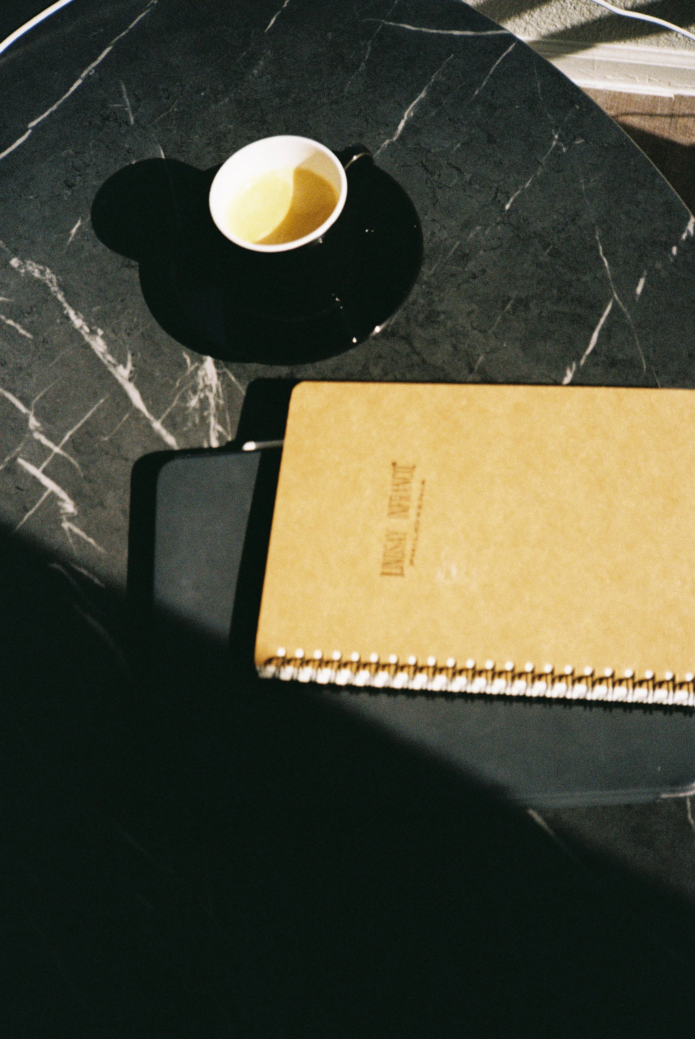 A black marble table with a white coffee cup casting a shadow, a yellow spiral-bound notebook titled 'DAYS PAPER' partially visible, and a smartphone lying underneath the notebook.