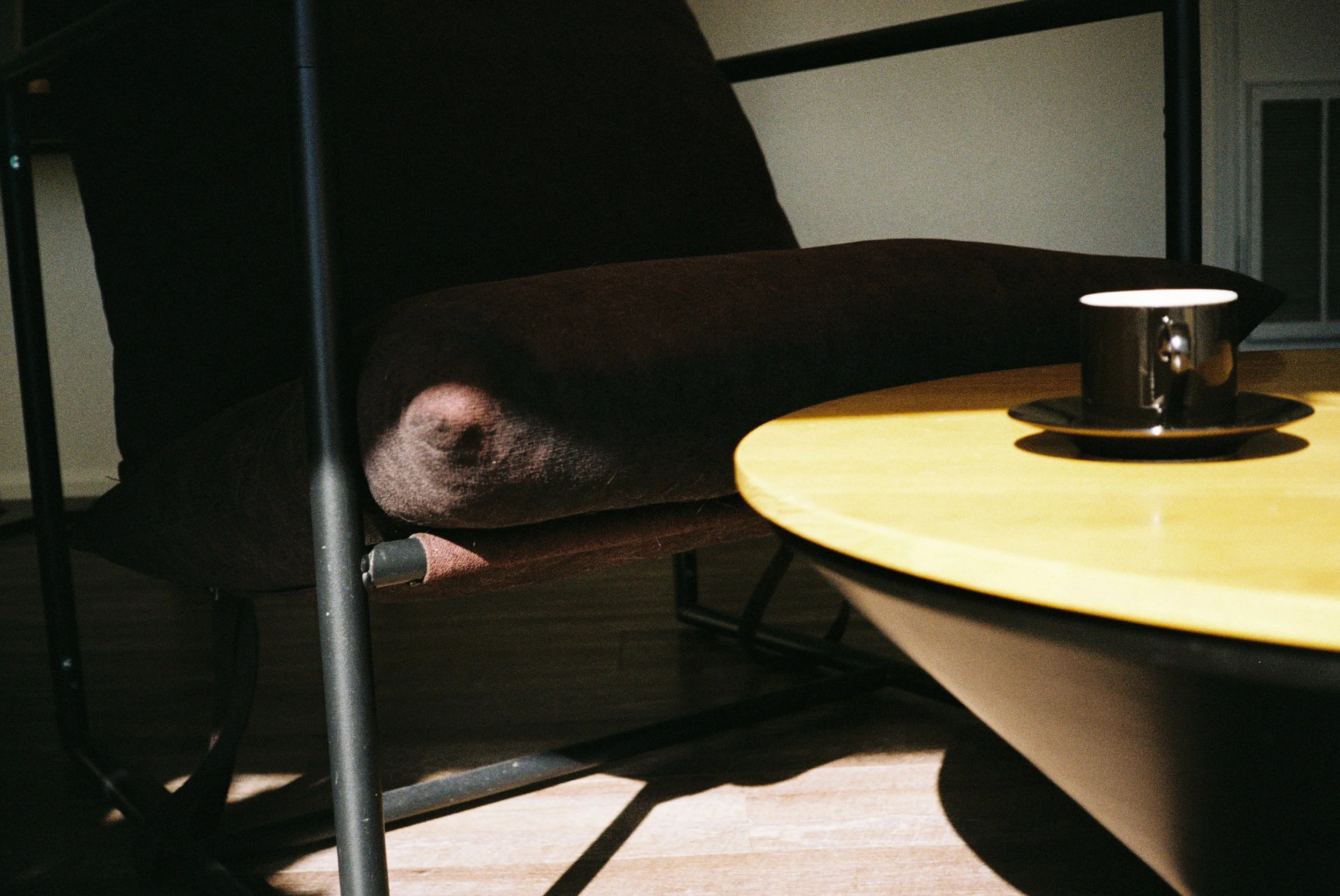 A brown cushioned chair with black metal frame beneath a wooden table, on which rests a black cup and saucer, with a partially visible window in the background.