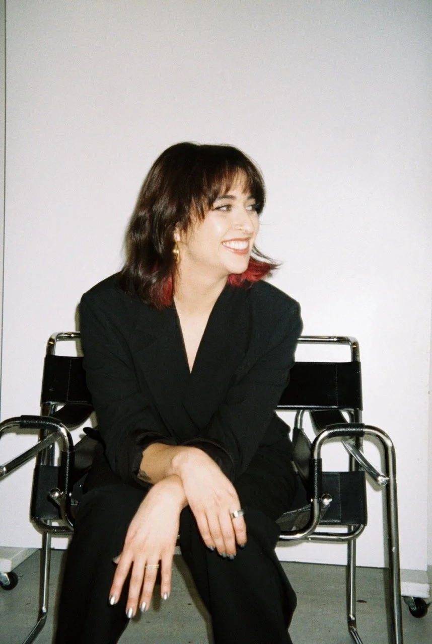 A woman with dark brown hair with red tips, wearing a black blazer, sitting on a black wheelchair against a plain white wall, smiling and looking to her right.