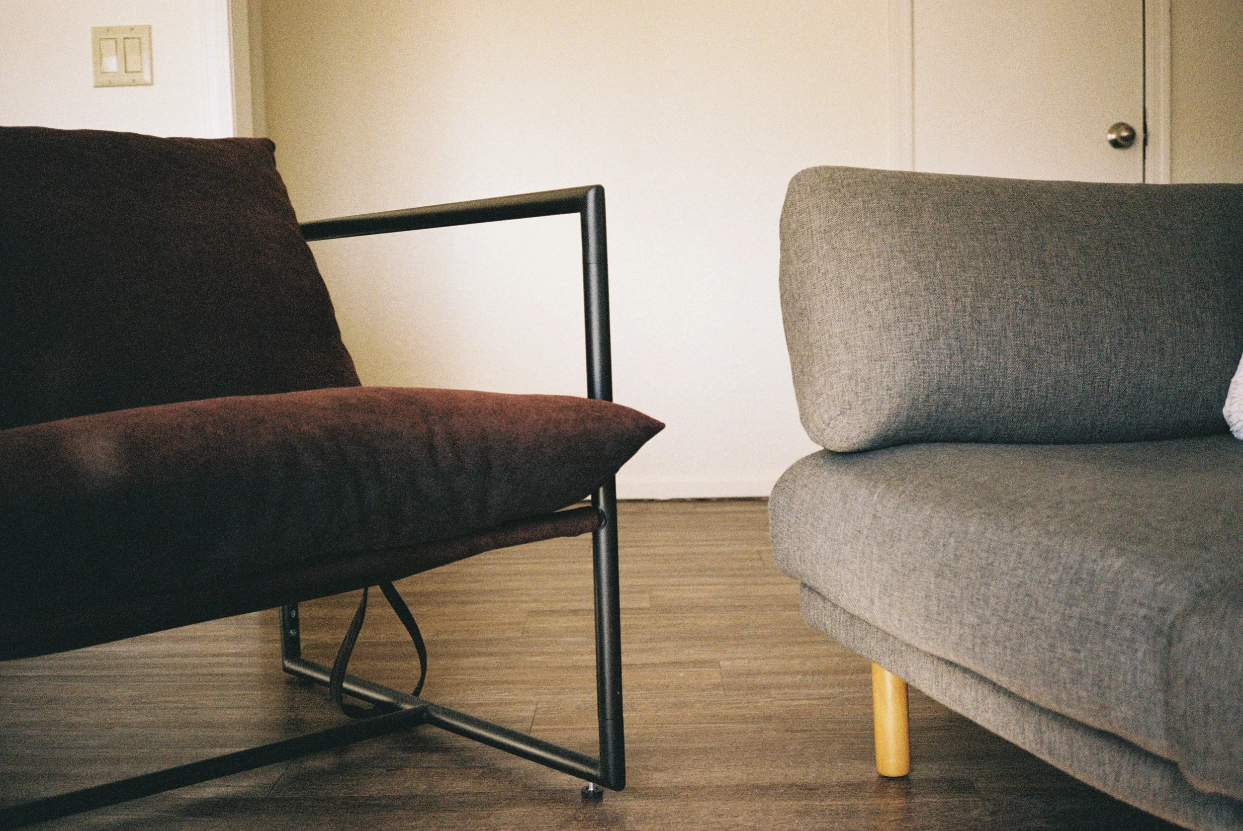 Interior of a room with two couches, one black with a metal frame and one gray with wooden legs, on a wooden floor with a plain wall and door in the background.