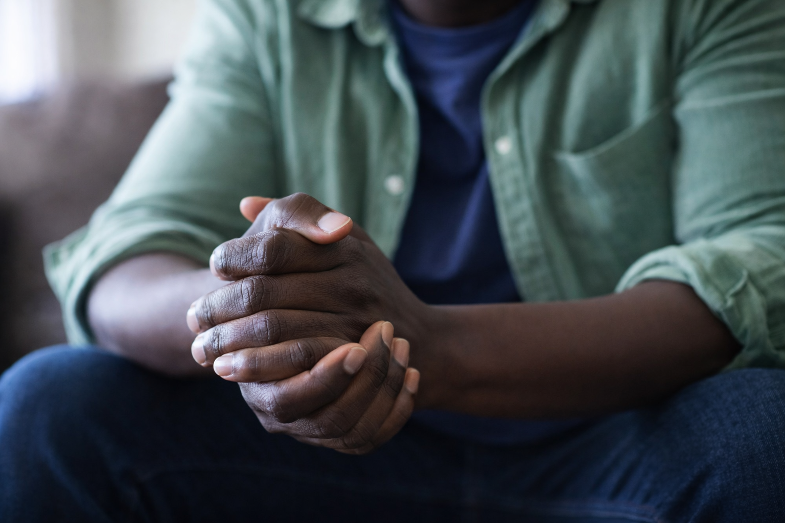 A person with dark skin sitting with their hands clasped together, wearing a green shirt over a blue t-shirt.