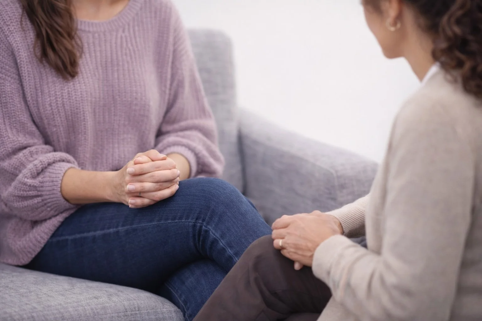 Two women sitting on a gray couch engaging in a conversation, one with hands clasped on her lap and the other with hands resting on her knee, in a well-lit room.