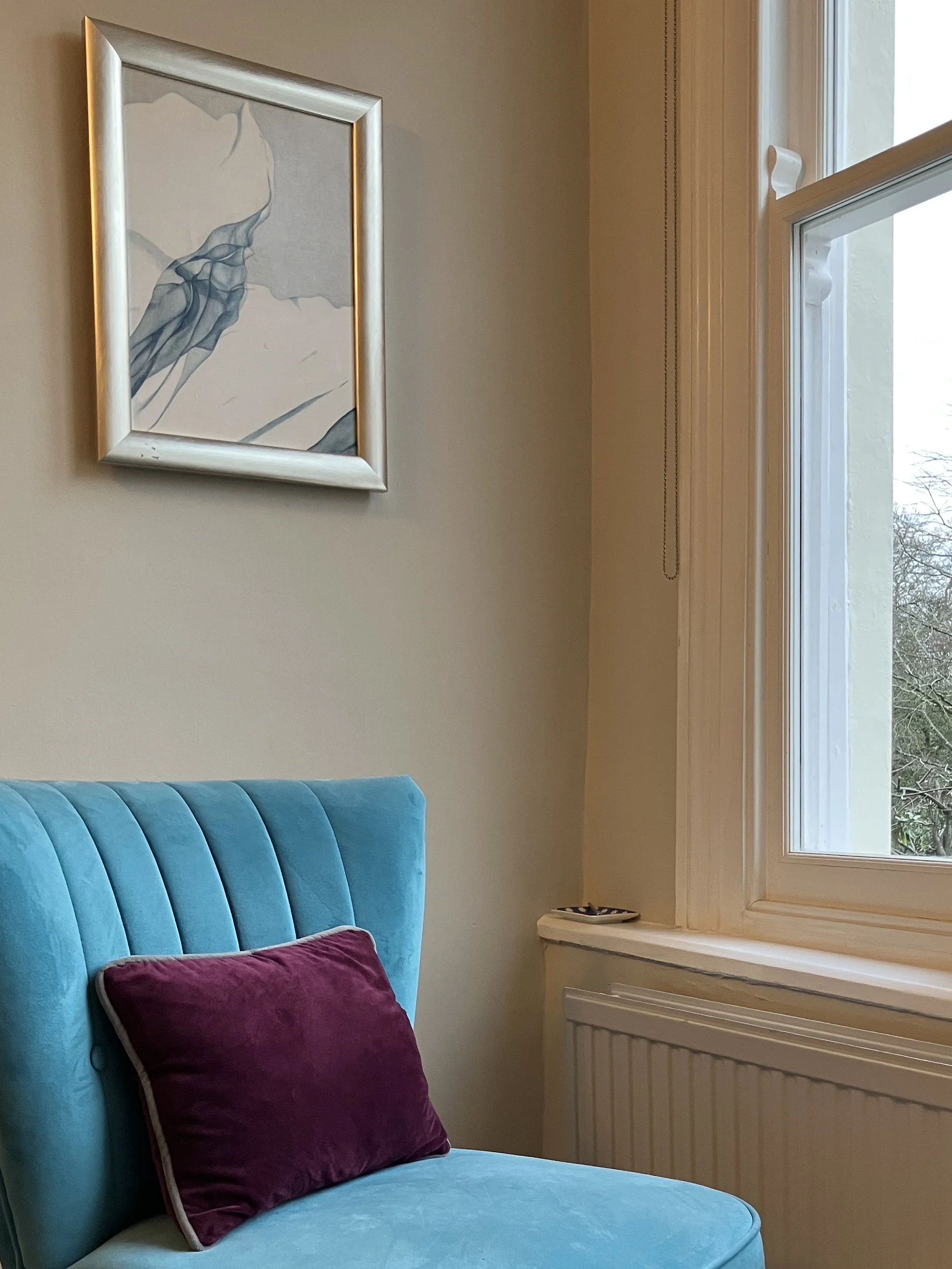 A corner of a room with beige walls, a large window with curtains, teal upholstered chair with vertical stripes, a purple velvet pillow on the chair, a decorative plate on the windowsill, and a framed abstract artwork hanging on the wall.