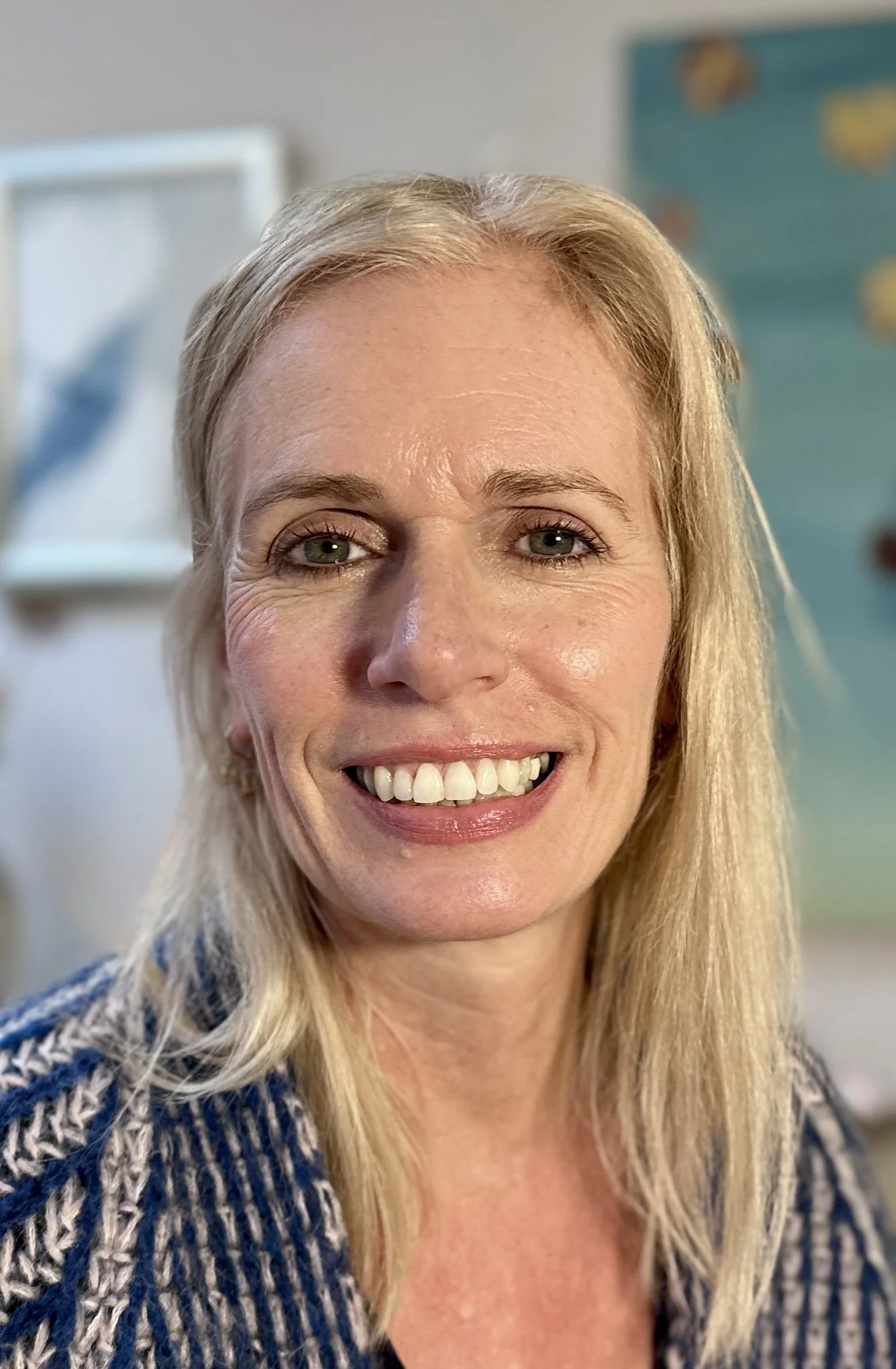 A smiling middle-aged woman with blonde hair and clear skin, wearing a patterned blue sweater, indoors with blurred artwork and pictures in the background.