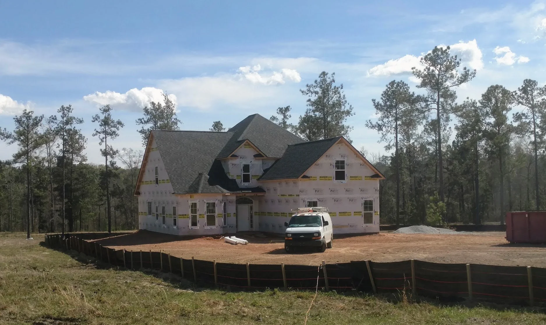 A large house under construction in a wooded area, with an unfinished exterior and a white construction van parked in front.