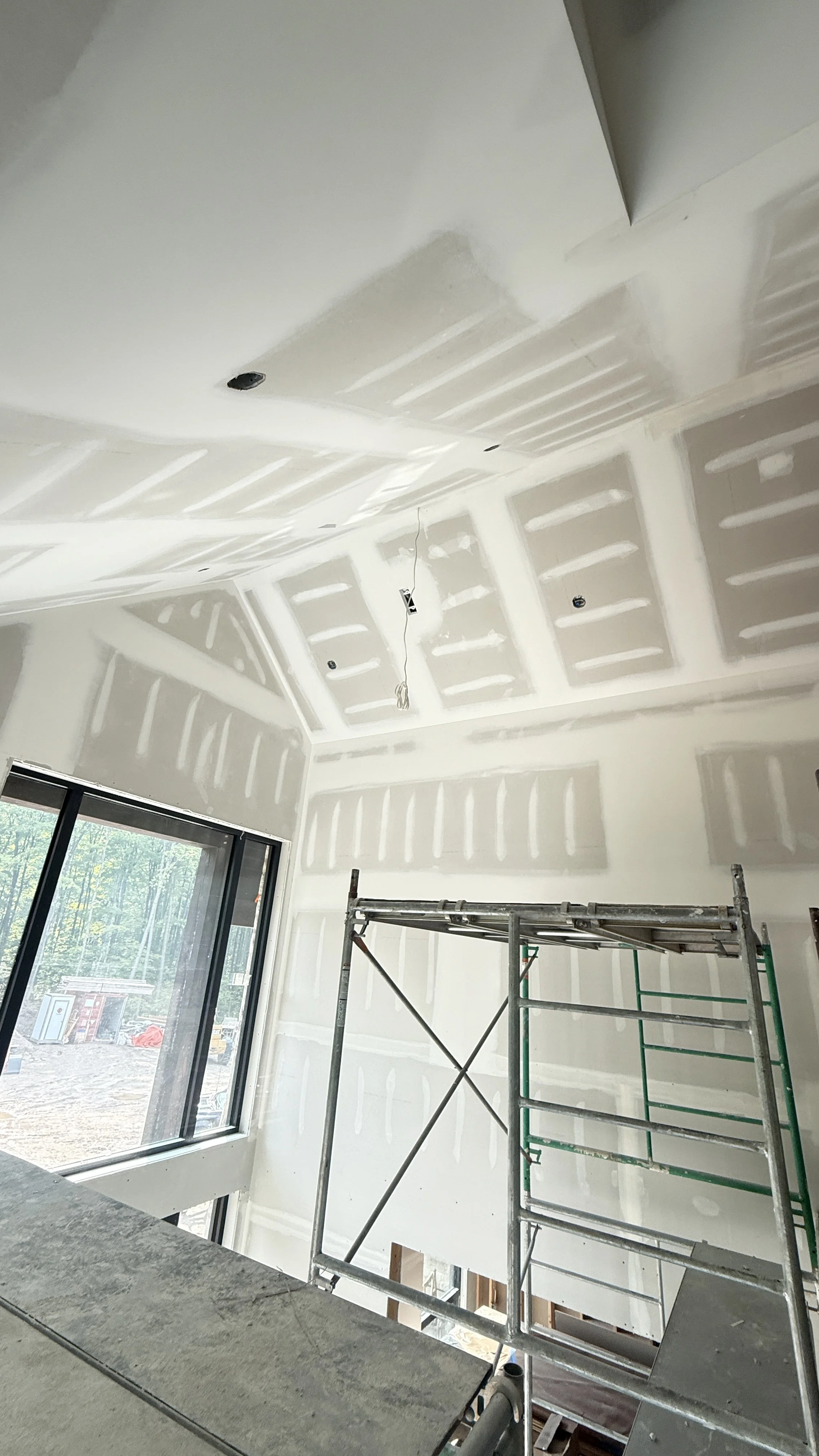 Interior of a house under construction with drywall installed on the walls and ceiling, scaffolding in the room, and a large window showing an outdoor construction area.