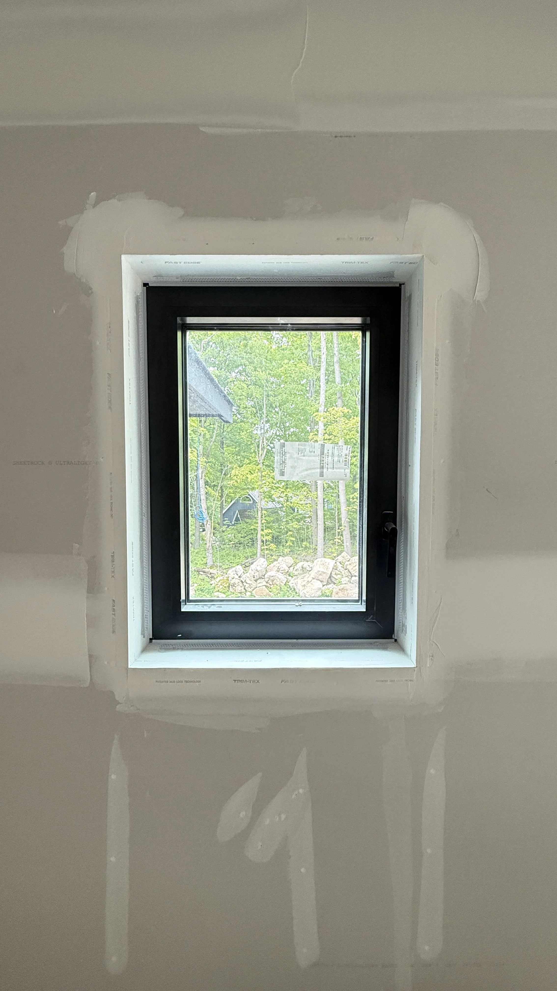 Interior view of a wall with a rectangular window, unfinished drywall around the window, and a view of trees and rocks outside.