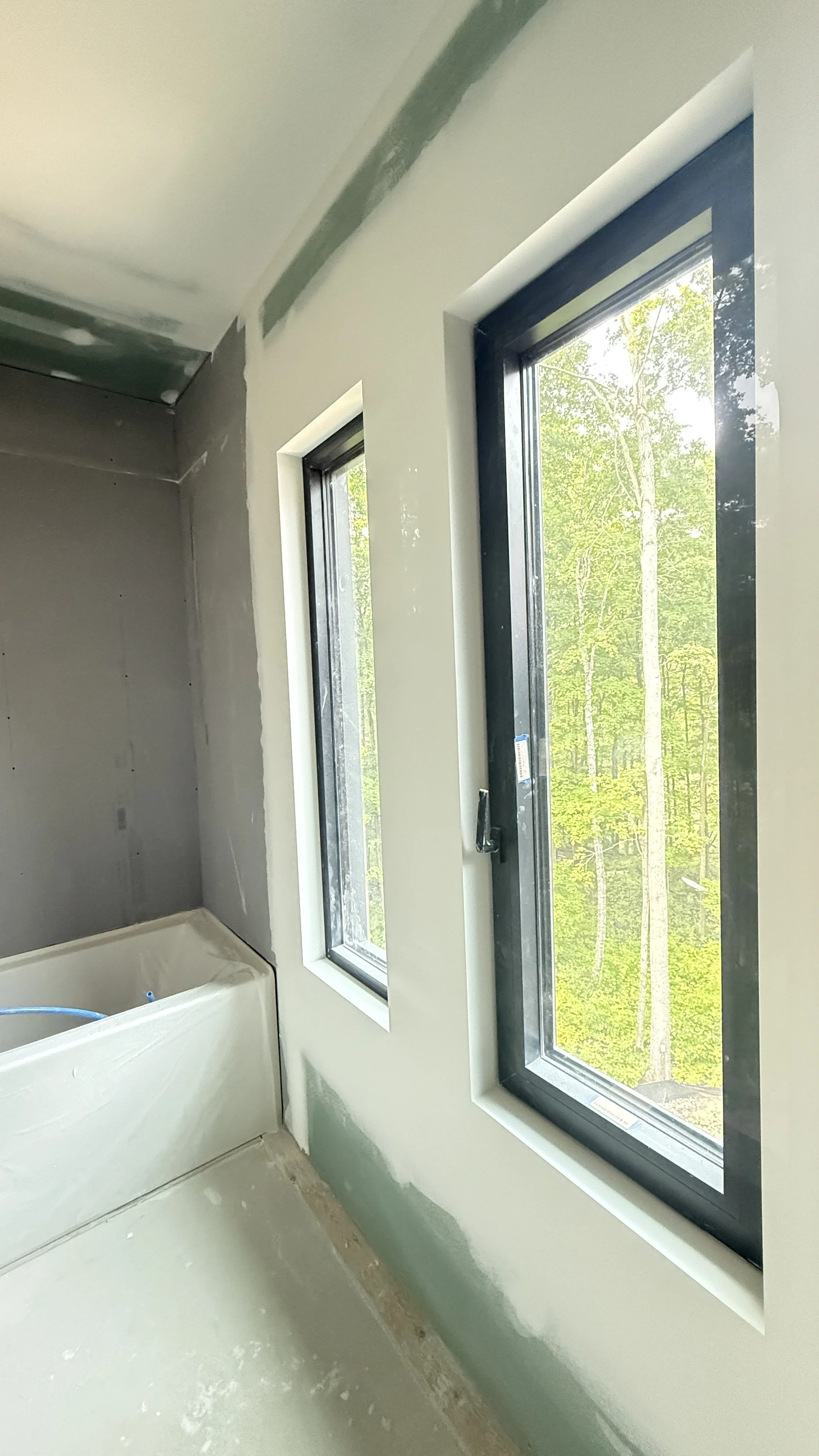 Interior of a room under construction, with two large windows and a gray drywall wall, showing unfinished edges and construction materials.