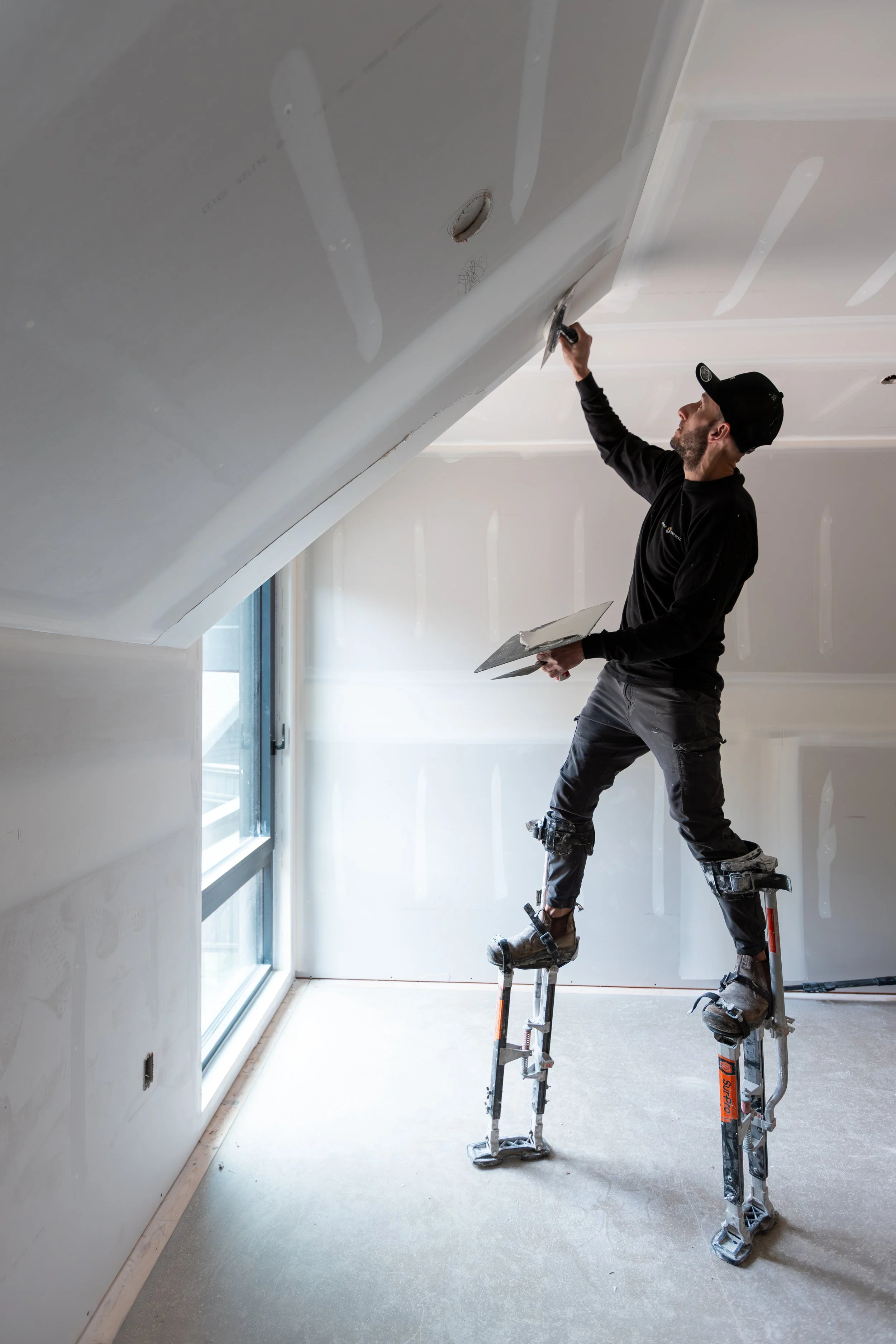A man working on drywall installation, standing on stilts, sanding the ceiling in a room with a sloped ceiling and a large window.
