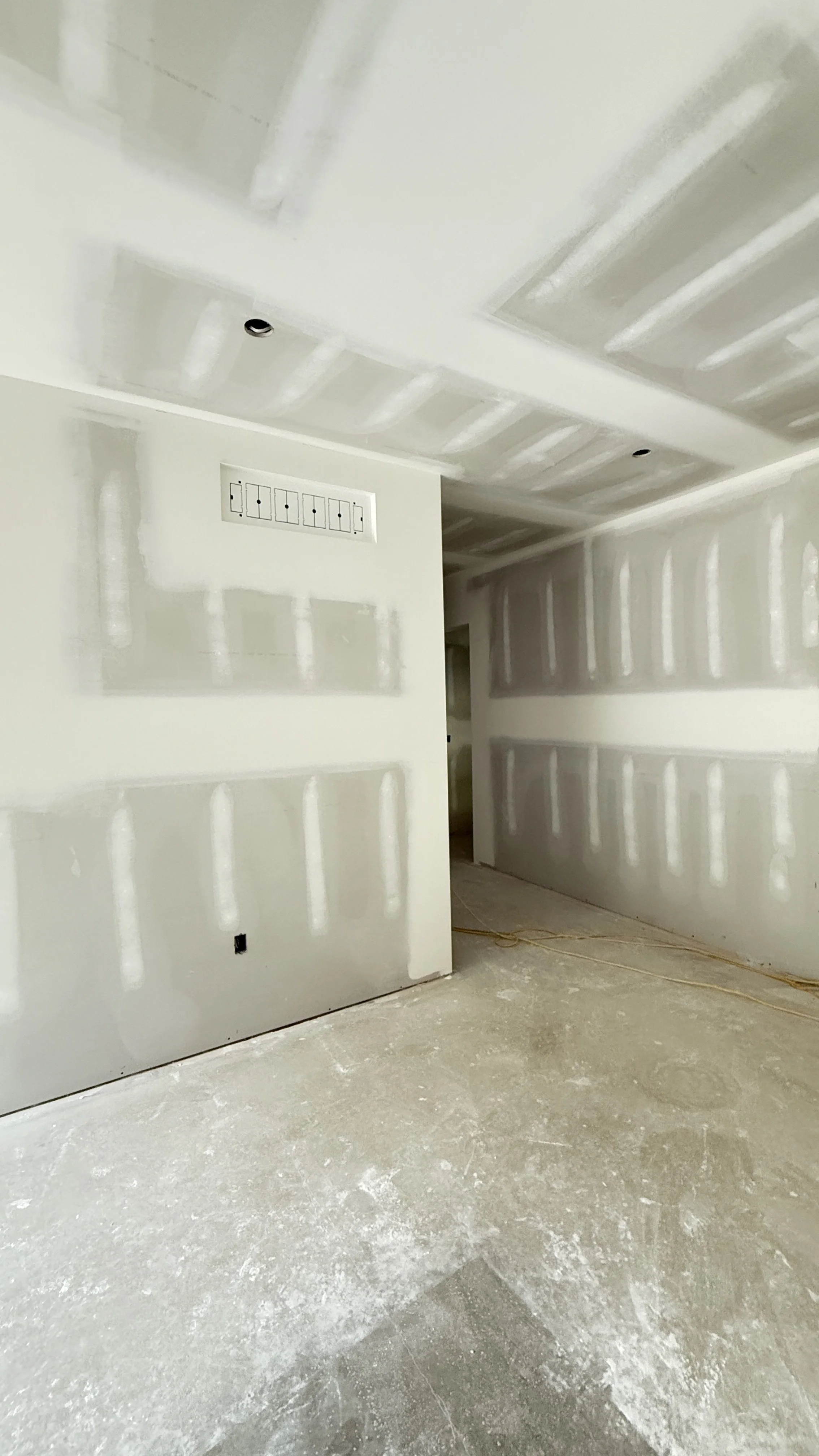 Interior view of a room under construction with drywall installed, including an electrical panel and visible wiring on the floor.