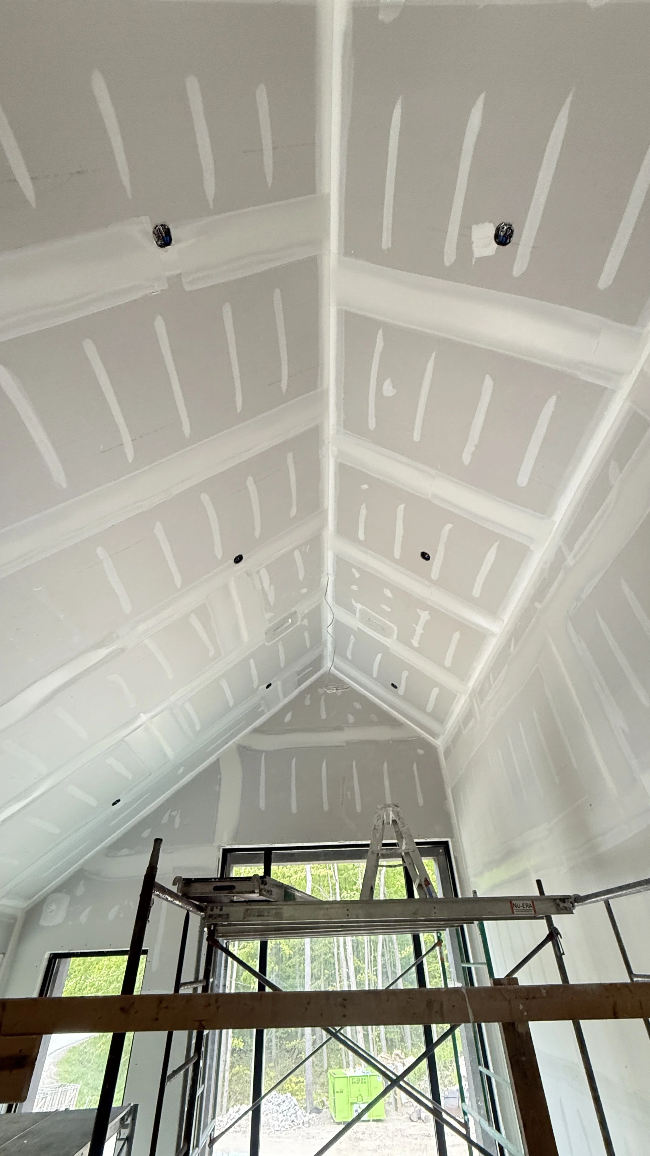 Interior view of a room under construction with drywall installed on the ceiling. Scaffolding and a ladder are present, and sunlight is coming through large window openings.