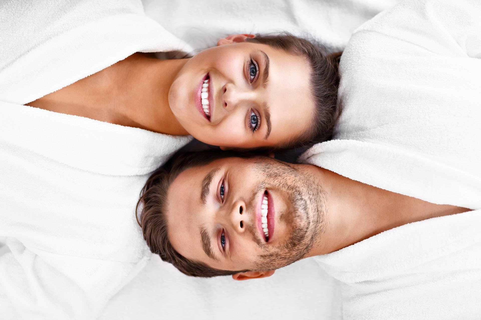 A smiling young woman and a smiling young man lying together under white blankets on a bed, facing the camera.