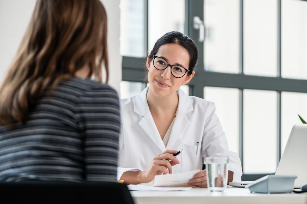 A woman with black hair and glasses, wearing a white coat, smiling and talking to another woman with reddish-brown hair and a striped shirt in an office setting.