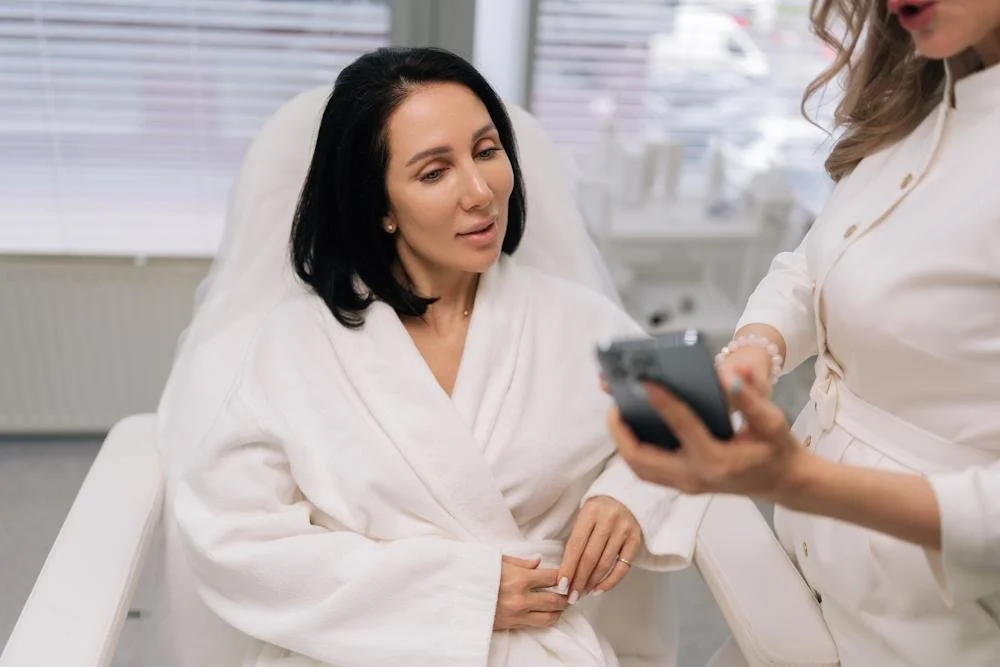 A woman in a white robe sitting on a medical examination chair, looking at a mobile phone held by another woman in a white coat, in a clinical setting.