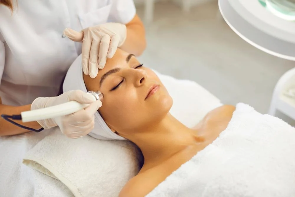 A woman receiving a facial treatment with a skincare device on her face in a spa or clinic setting, while lying down with her eyes closed.