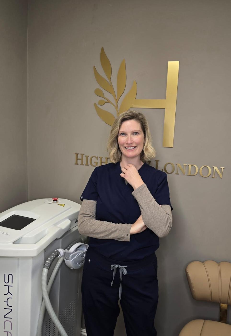 A woman with blonde hair smiling, wearing navy medical scrubs with a beige long-sleeve shirt underneath, standing next to a medical device in a room with a sign that reads 'Highland London' on the wall.