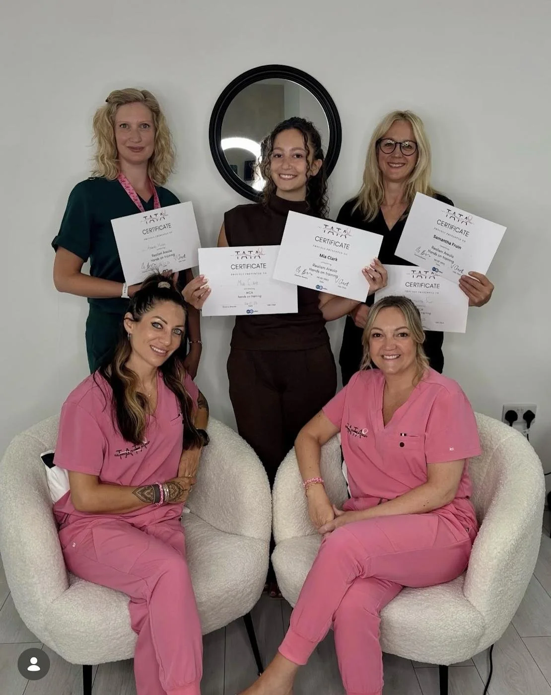 Six women, some in pink scrubs and some in dark tops, holding certificates, posing in a room with a white wall and a round black mirror behind them.