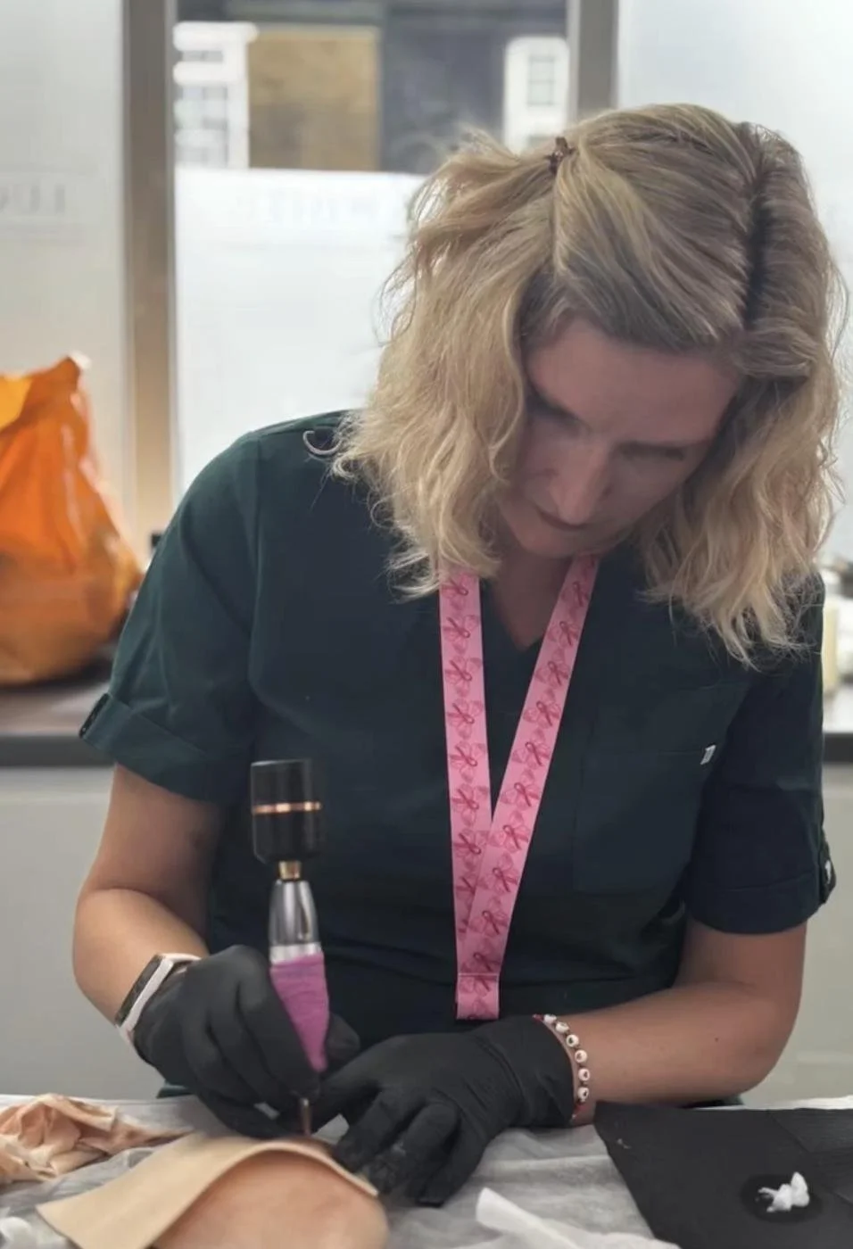 A woman with blonde curly hair wearing black gloves and a pink lanyard uses a tattoo machine on a person's skin in a well-lit room.