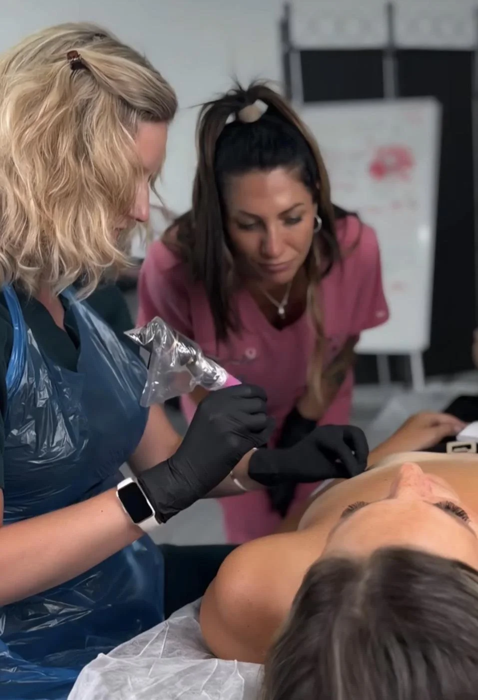 Two healthcare professionals performing a cosmetic procedure on a patient lying down in a clinical setting.