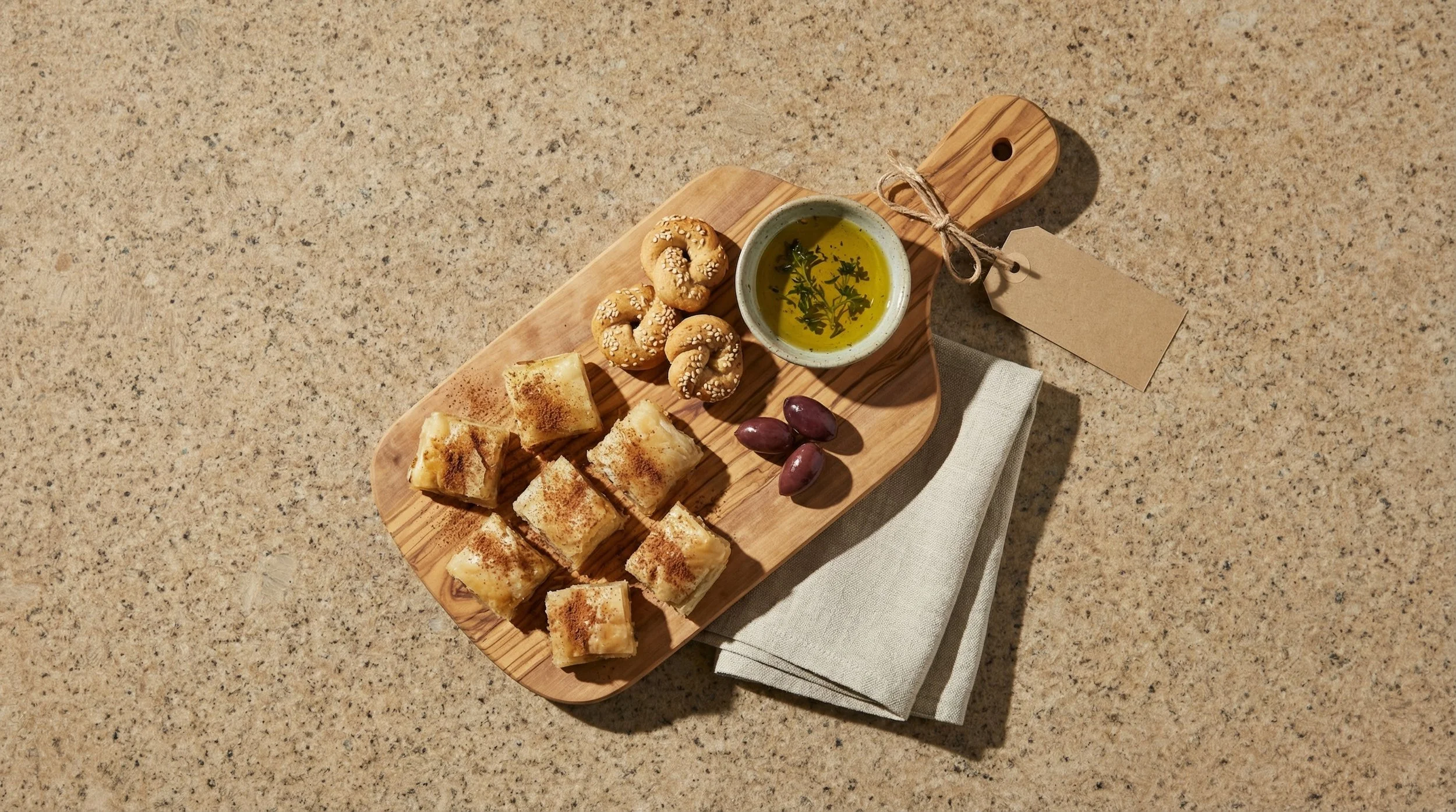A wooden serving board with small cheese pastries, purple grapes, a bowl of olive oil with herbs, and sesame seed crackers, placed on a beige countertop with a white cloth underneath.