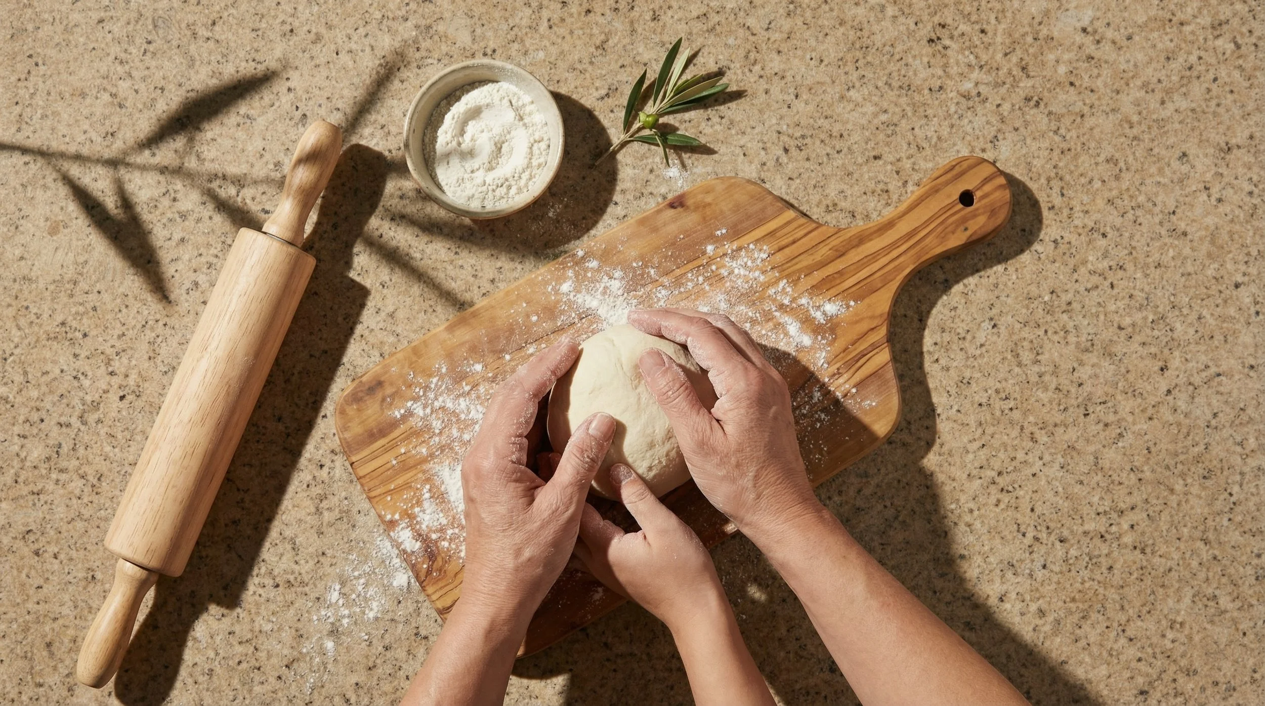 Hands kneading dough on a wooden cutting board, with a rolling pin, a bowl of flour, and a small plant on a sandy-colored countertop.