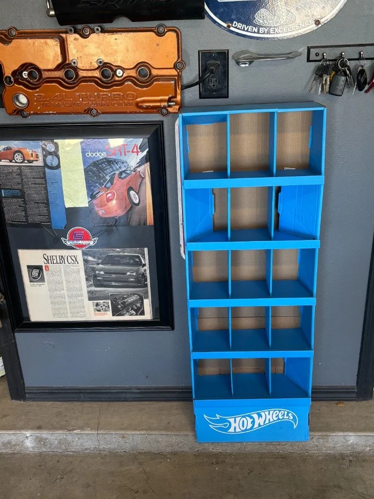 Blue Hot Wheels display shelf with six open compartments against a gray wall, with automotive posters, a valve cover, and a hook with keys nearby.
