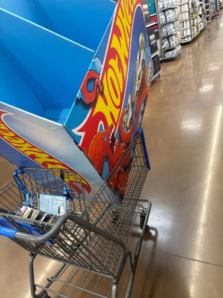 A large Hot Wheels display box in a supermarket shopping cart, with the box featuring colorful graphics of cartoon dinosaurs and cars.