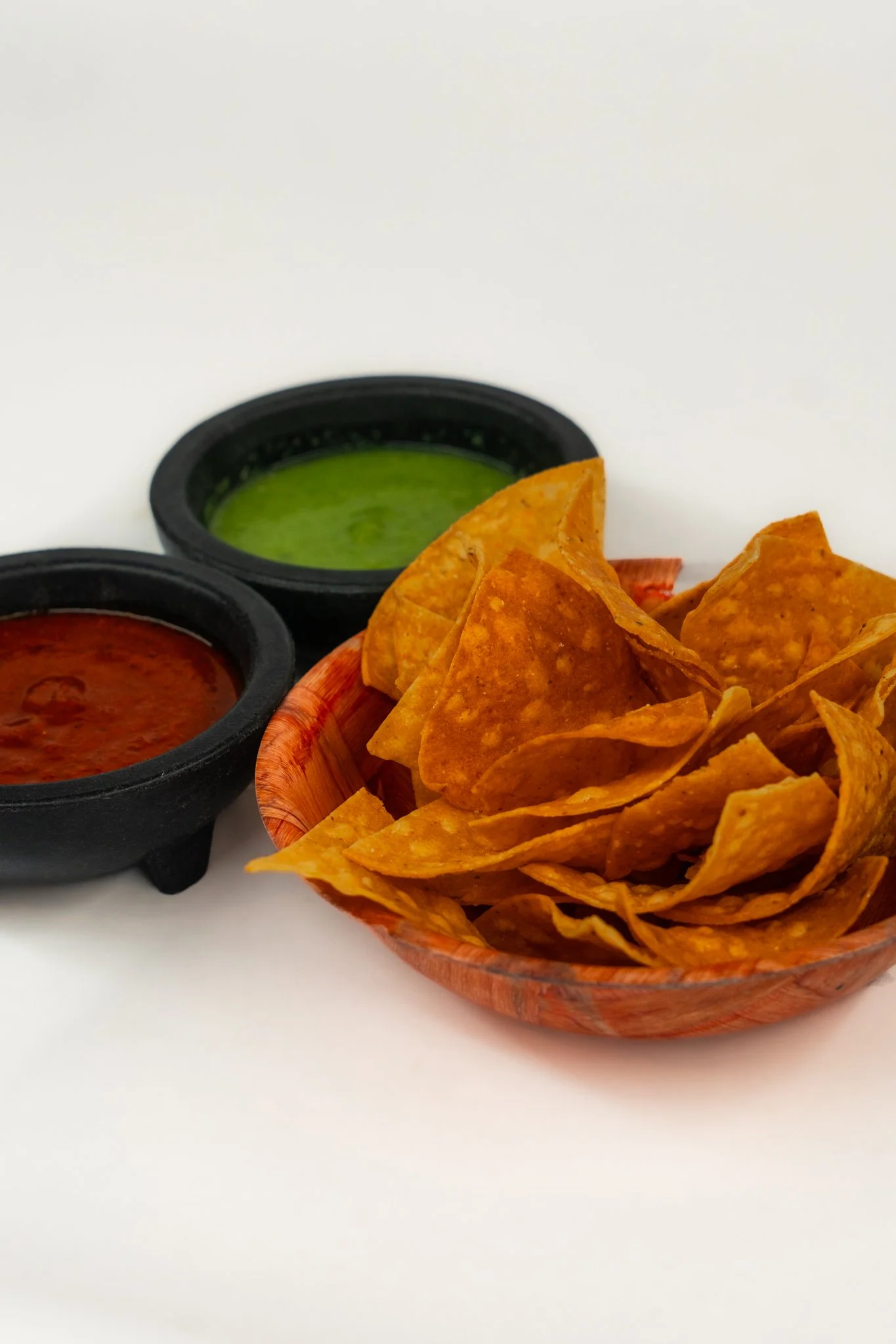 A wooden bowl filled with crispy tortilla chips next to two small black bowls of red and green salsa on a white surface.