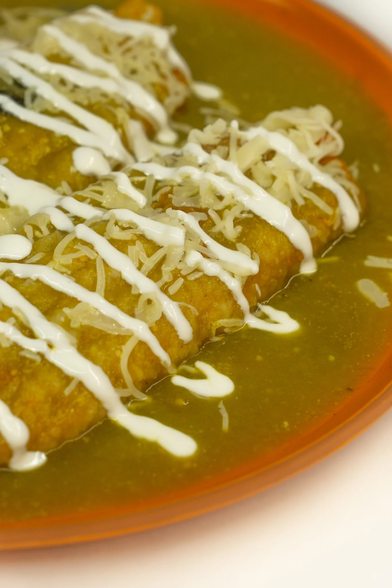 Fried cheese enchiladas topped with shredded cheese and drizzled with white sauce, served in green enchilada sauce.
