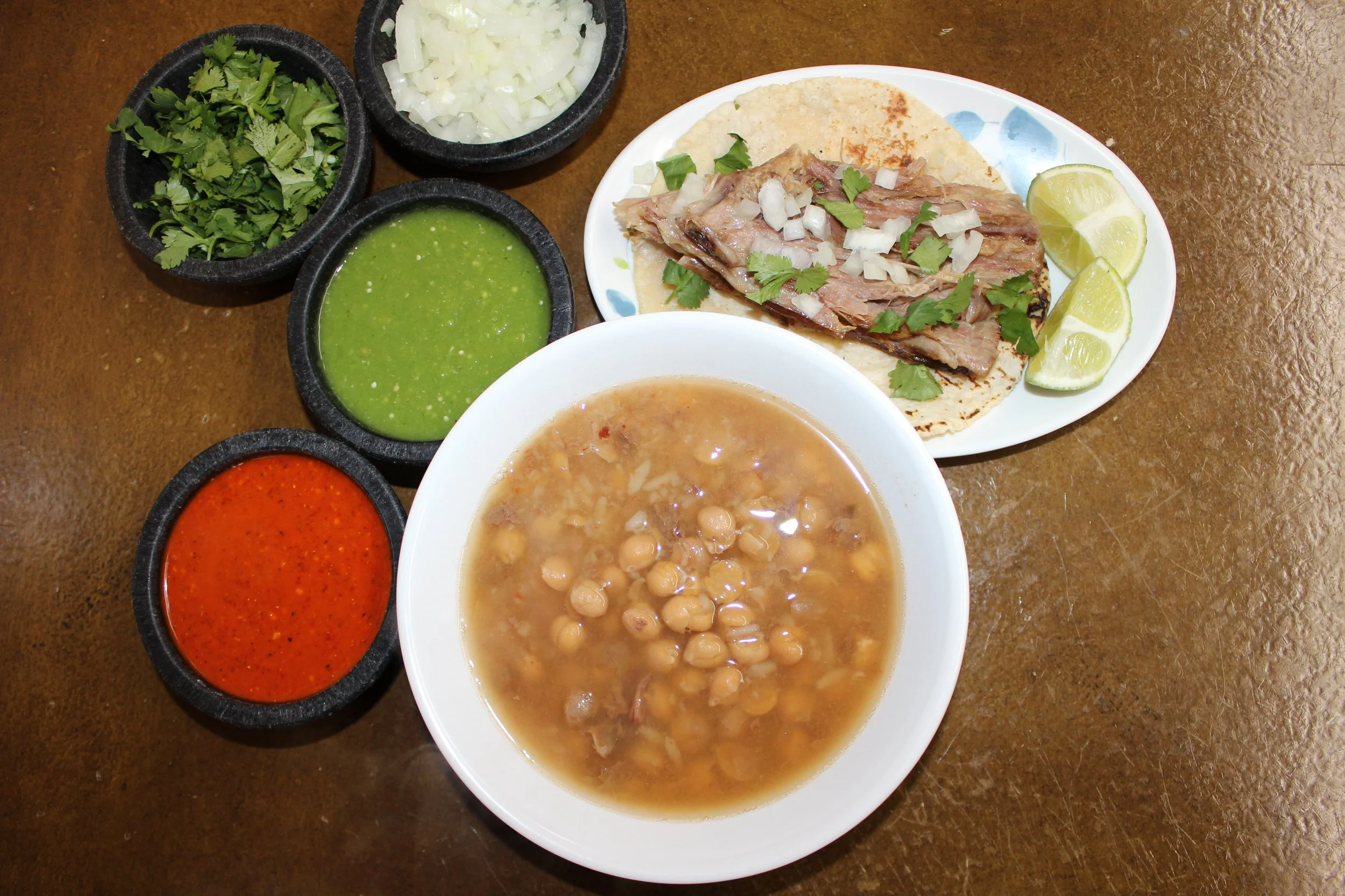 Traditional Mexican meal with a bowl of chickpea soup, corn tortillas with sliced grilled beef topped with onions and cilantro, and small bowls of chopped onions, cilantro, salsa verde, and salsa rojo, all on a wooden table.