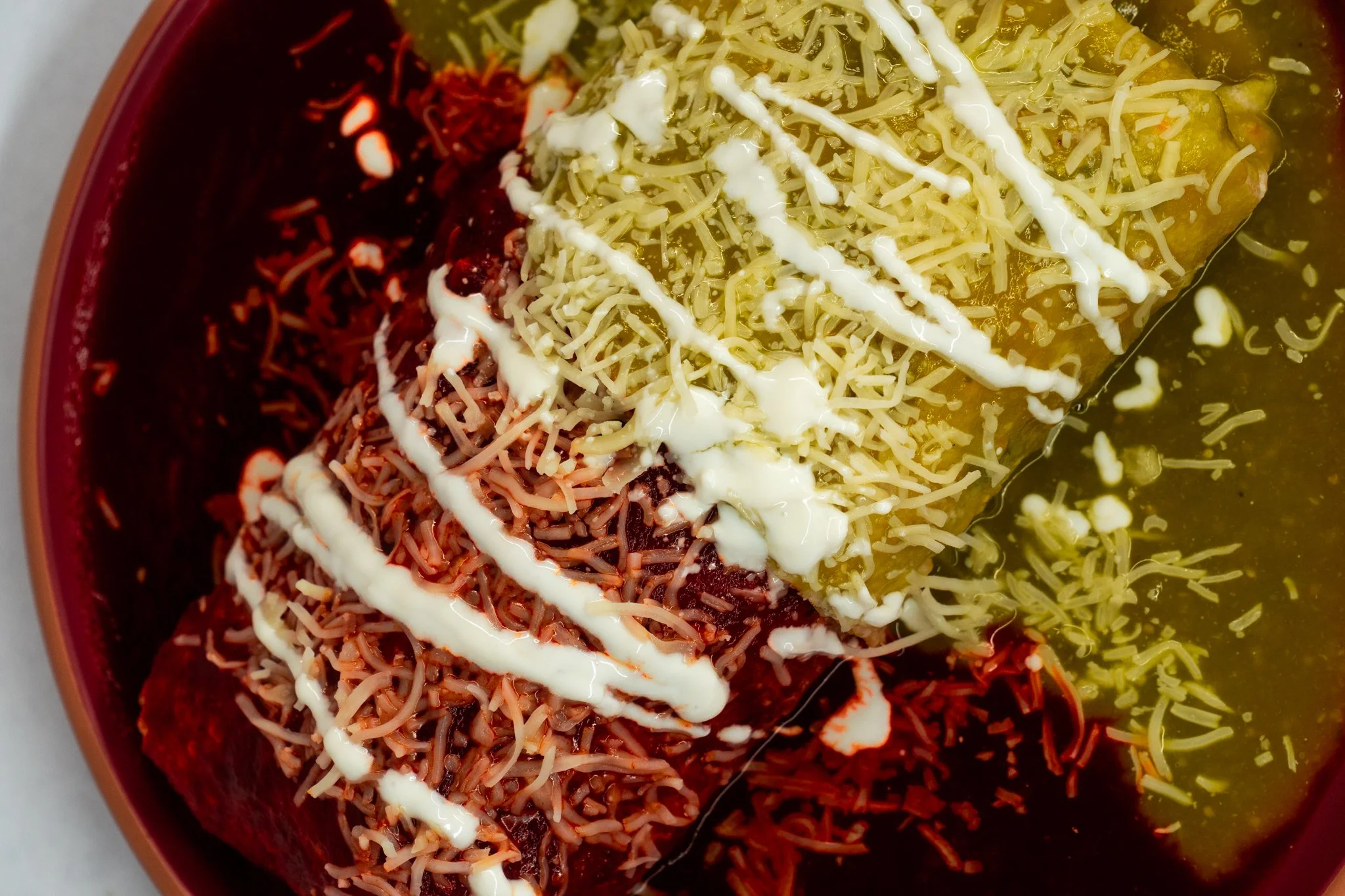 A colorful plate of Mexican food featuring enchiladas topped with shredded cheese, sour cream, and a drizzle of white sauce, with a side of green sauce.