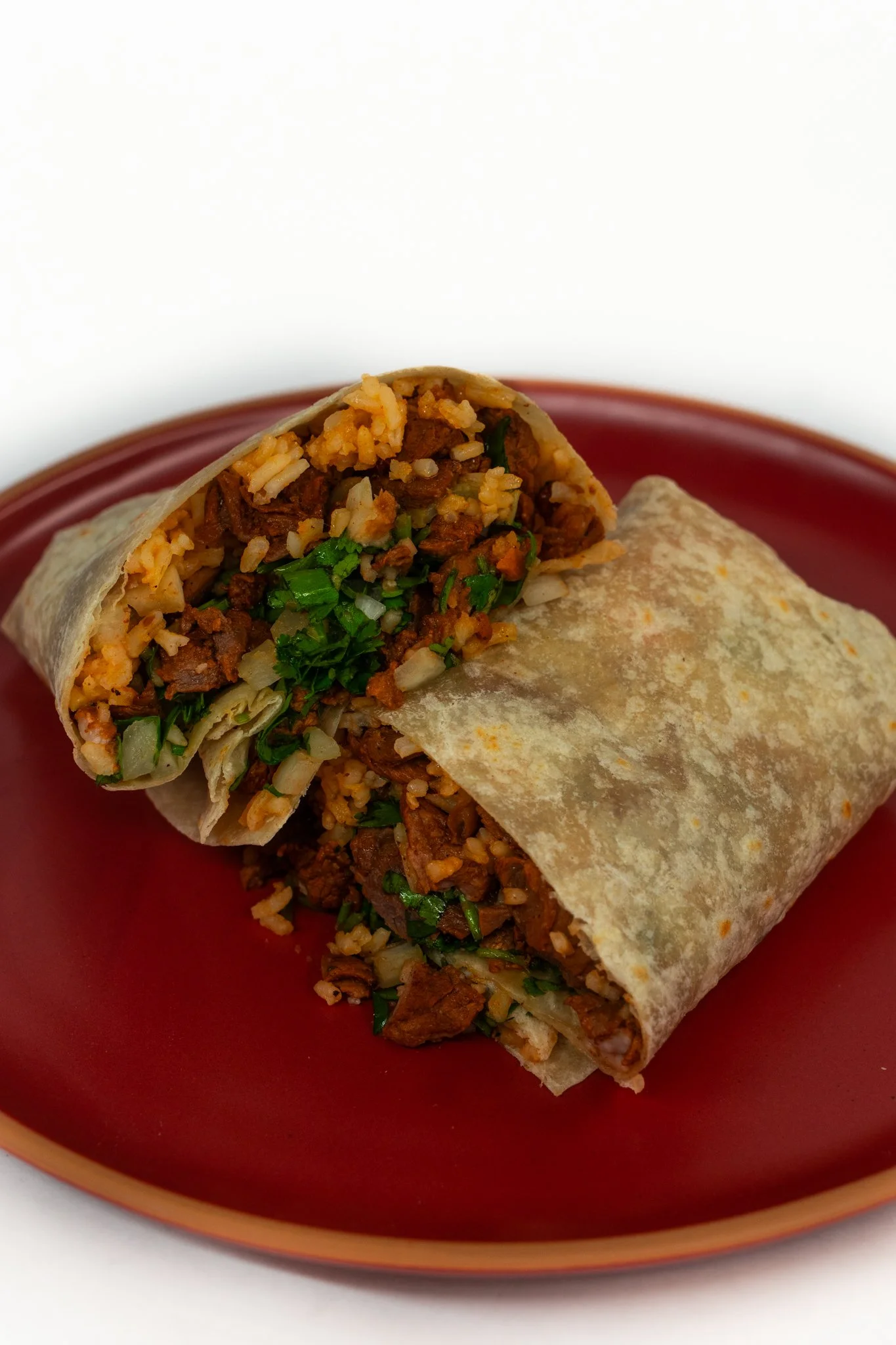 A burrito cut in half, showing seasoned ground beef, rice, beans, chopped onions, and chopped cilantro inside a flour tortilla on a red plate.