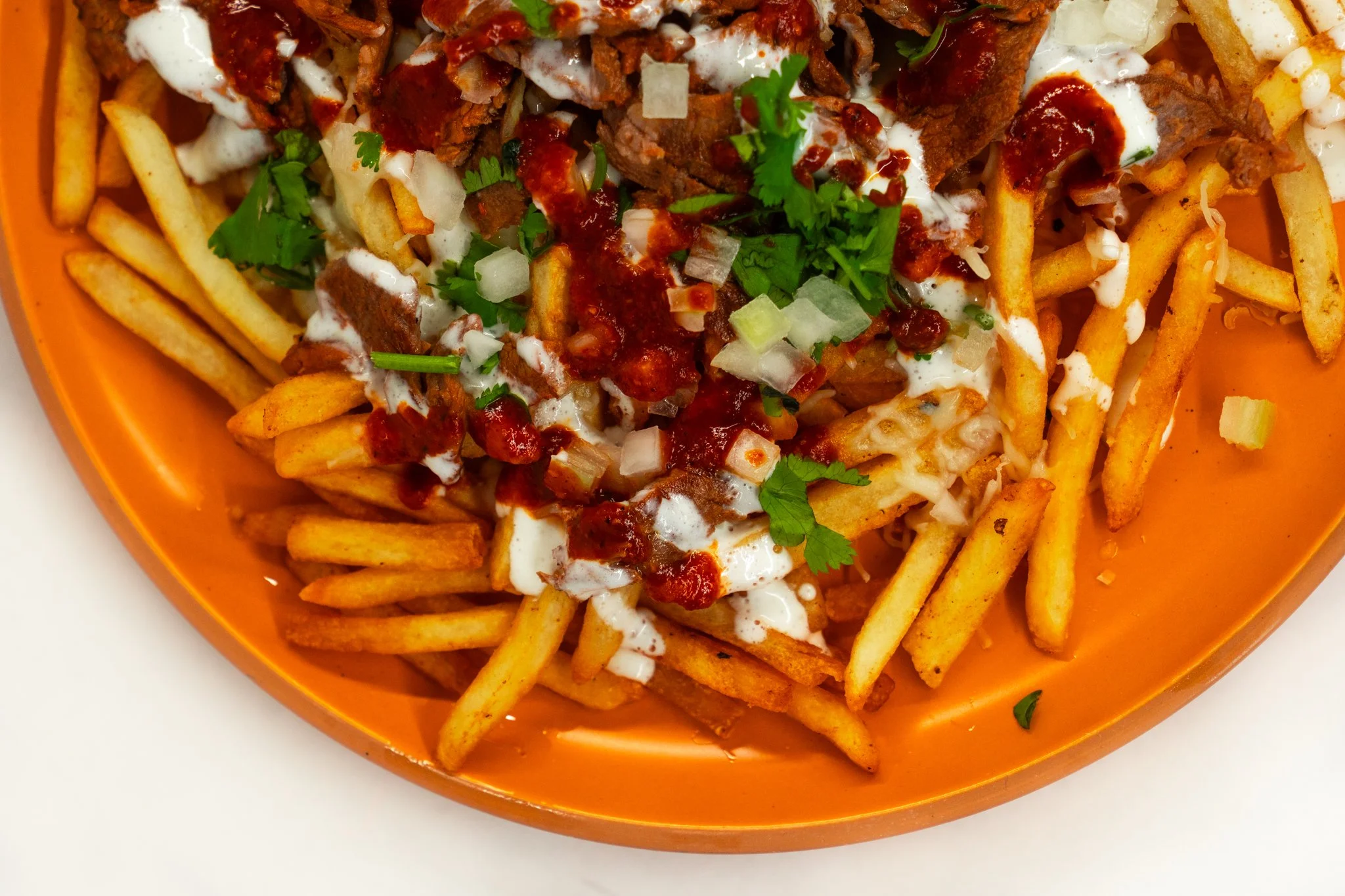 French fries topped with shredded cheese, chopped onions, cilantro, and drizzled with sour cream and hot sauce in a bright orange serving dish.