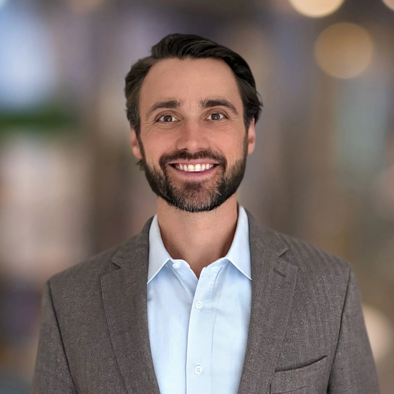 Portrait of a smiling man with brown hair, a beard, wearing a white shirt and gray blazer, against a blurred background with warm lighting.