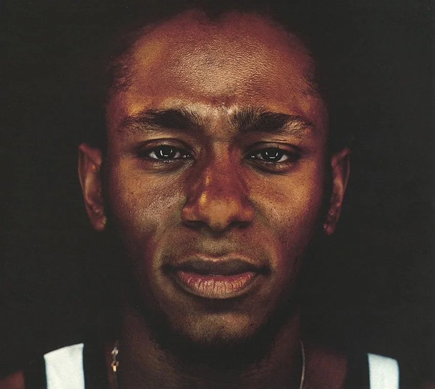 Close-up portrait of a young man with dark skin, short hair, and wearing a black and white sleeveless shirt. The background is black.
