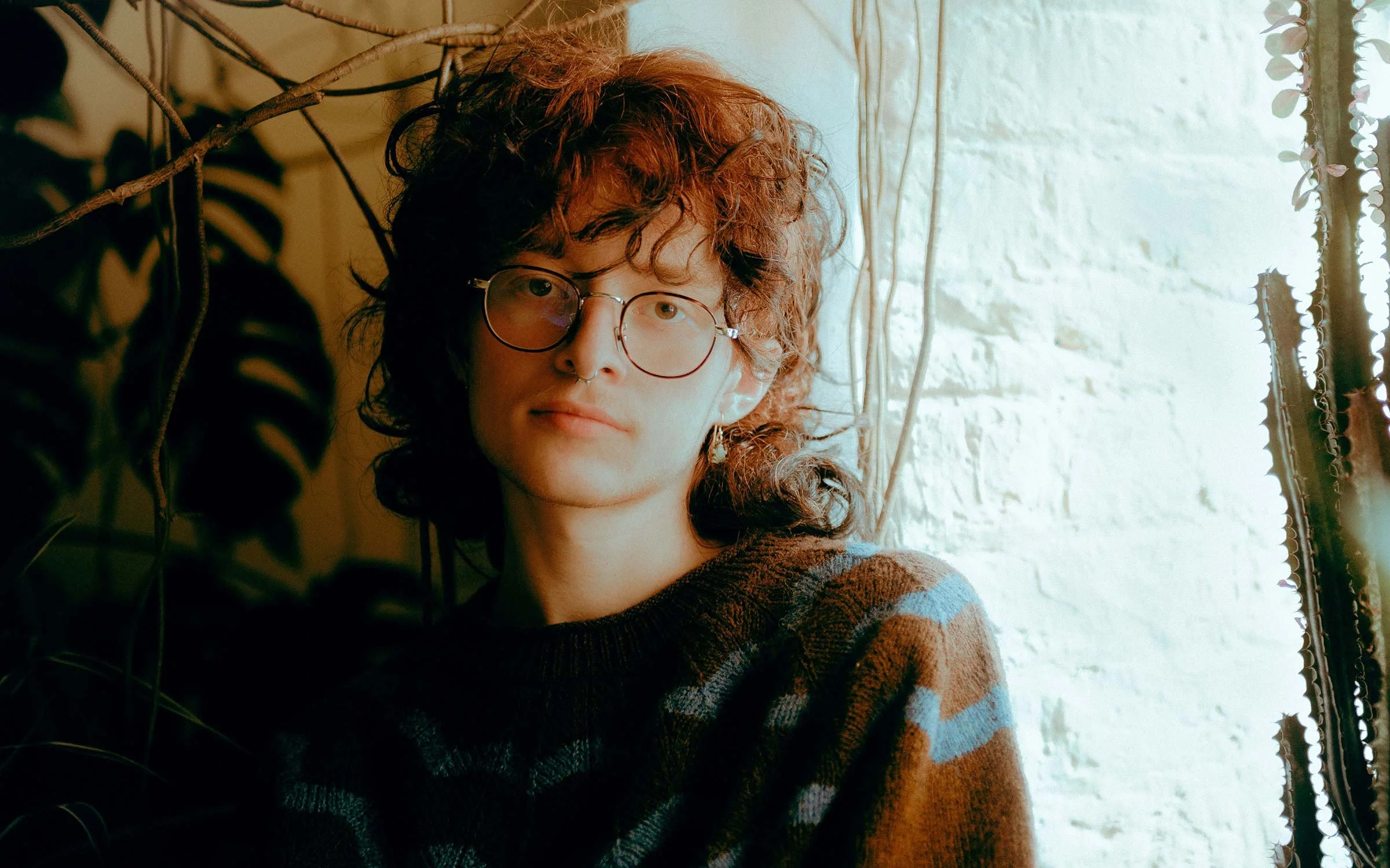 A young person with curly reddish hair and glasses, wearing a patterned sweater, leaning against a white brick wall near a window with sunlight, and surrounded by houseplants.