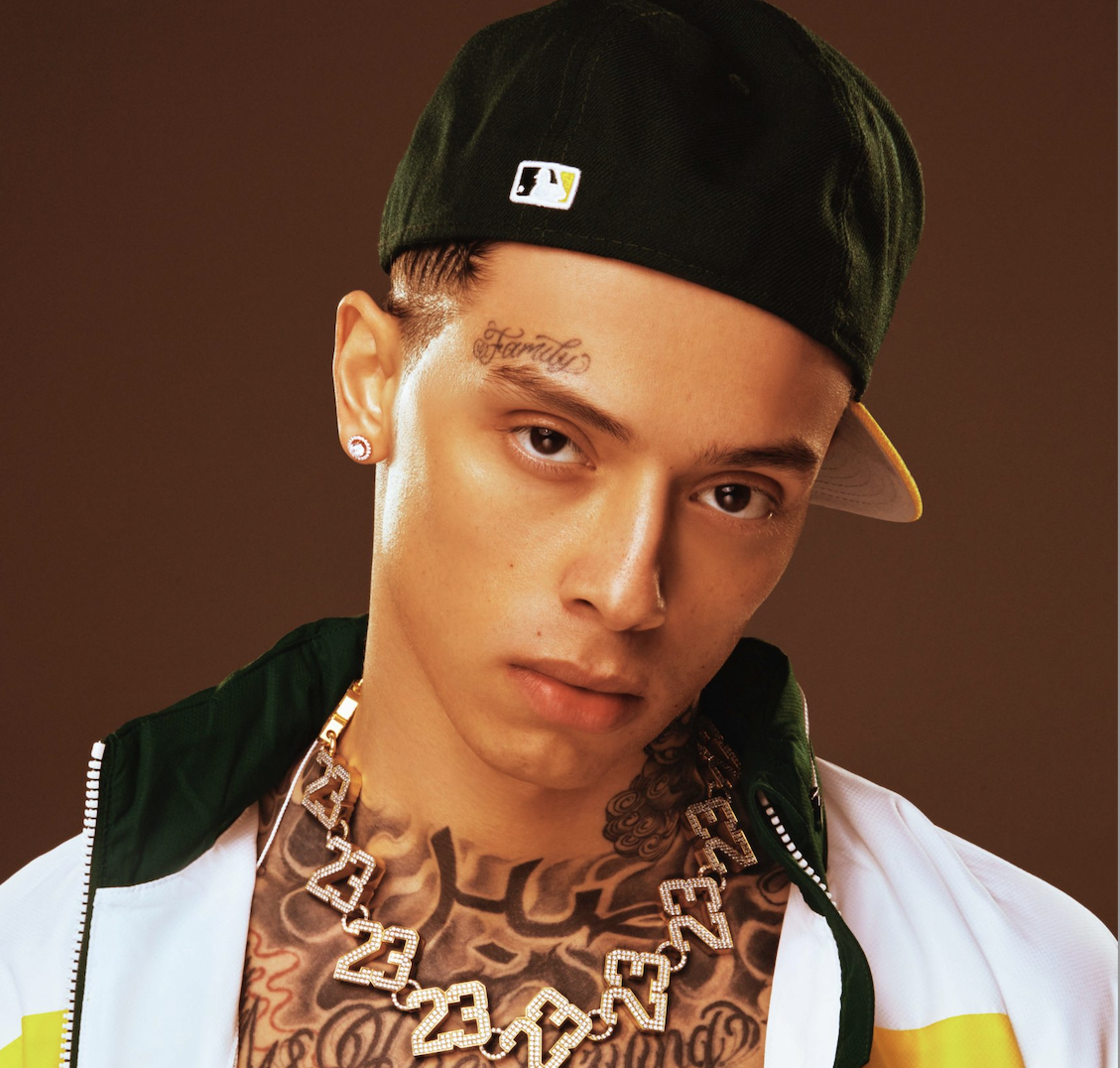 Young man with tattoos, wearing a black baseball cap, diamond necklace, and earrings, posing against a brown background.