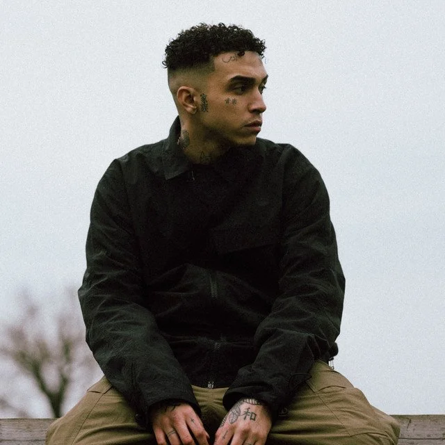 Young man with tattoos, short curly hair, wearing a black jacket and tan pants, sitting outdoors against a cloudy sky and blurred background.