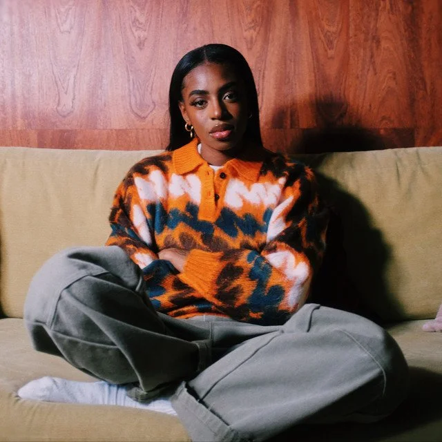 A woman with dark hair, wearing a colorful orange, blue, black, and white sweater, leather pants, and white socks, sitting on a beige couch with a wooden wall background.
