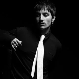 Black and white portrait of a young man with short dark hair, wearing a dark shirt and a light-colored tie, against a dark background.