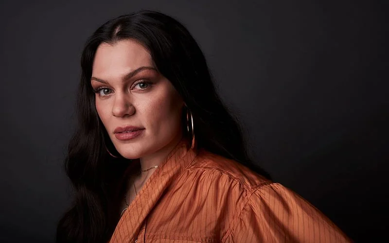 A woman with long dark hair, wearing large hoop earrings and an orange blouse, posing against a dark background.