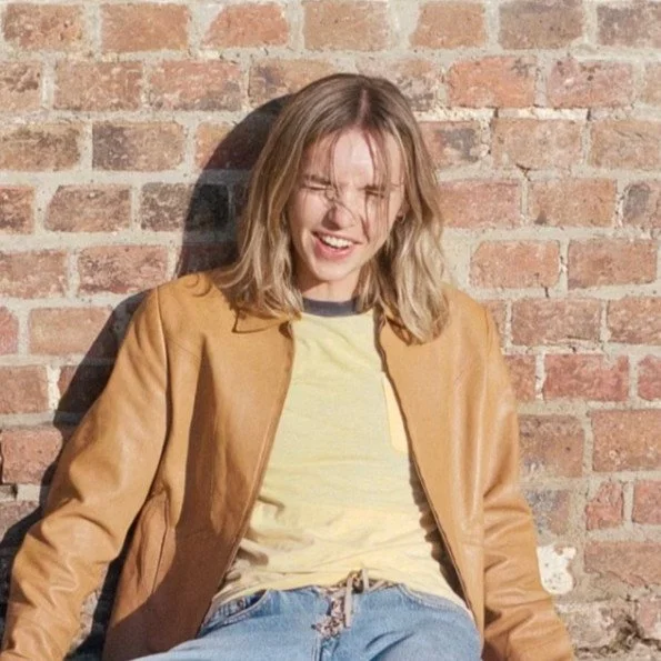 A woman with shoulder-length blonde hair smiling and squinting, sitting against a brick wall, wearing a tan jacket, yellow t-shirt, and blue jeans.