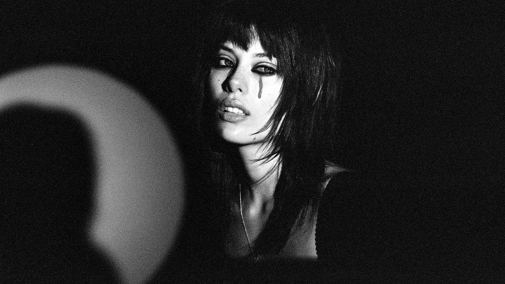 Black and white photo of a woman with messy dark hair, partially covered face, with one eye slightly closed, showing a tear on her cheek, in a dark background.