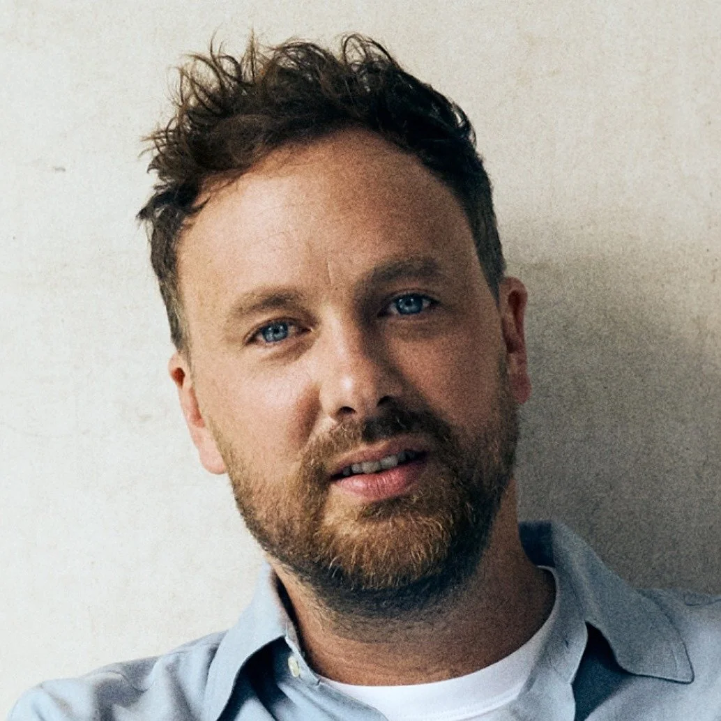 Close-up of a man with blue eyes, curly brown hair, and a beard, wearing a light blue collared shirt, standing against a plain wall.