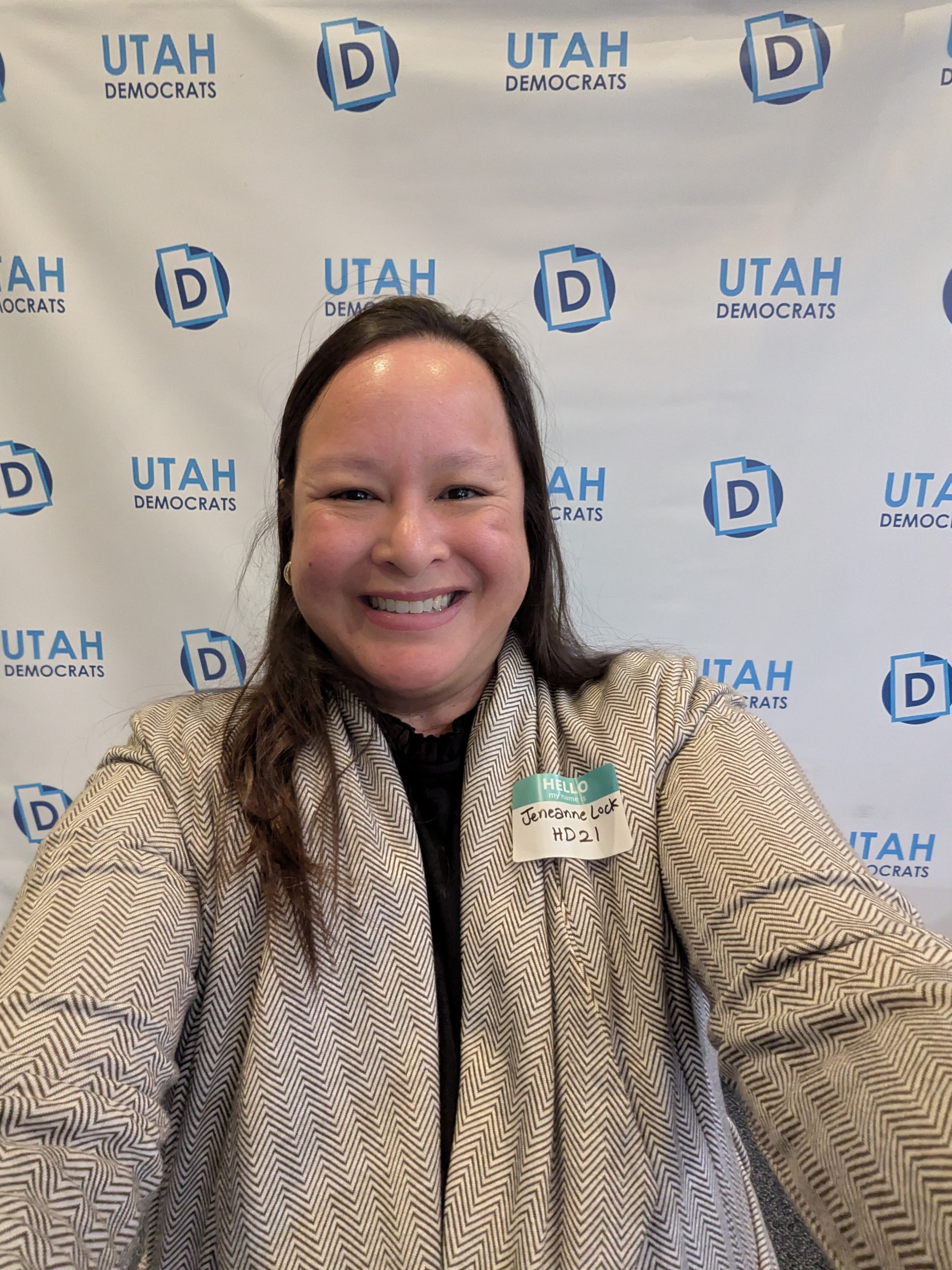 A woman taking a selfie at an event with a Utah Democrats backdrop. She is smiling, wearing a beige zigzag blazer, and has a name tag that reads 'Jeneanne Louw' and 'HD21'.