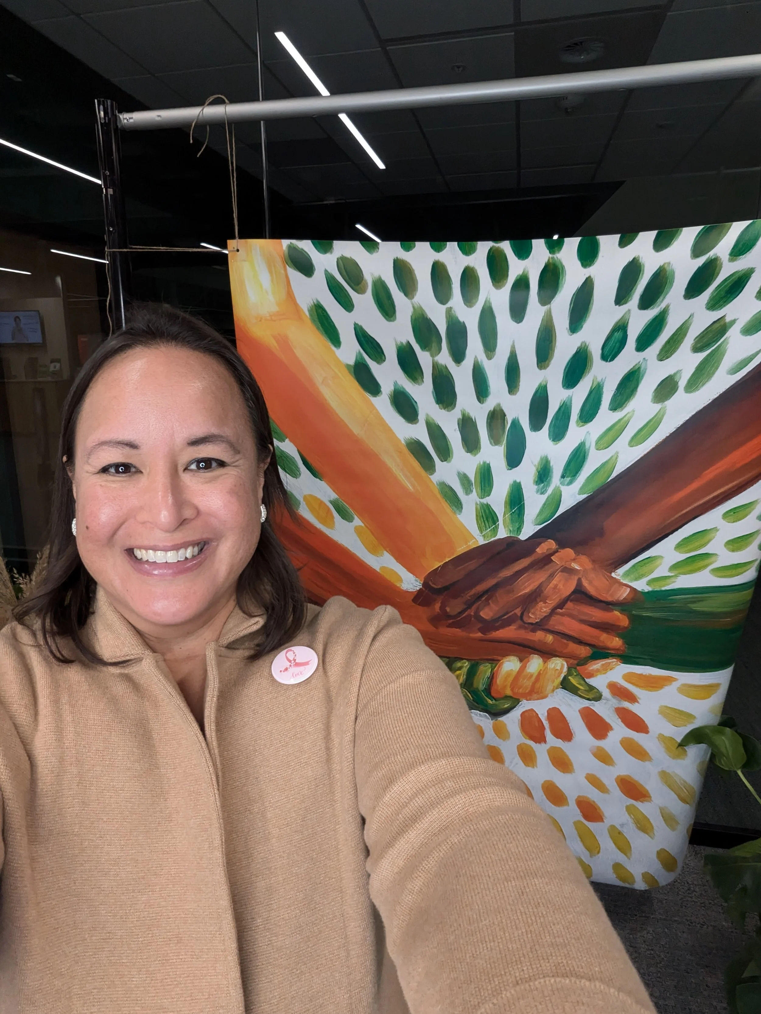 A woman taking a selfie in front of a colorful artwork depicting two hands shaking with brushstrokes of green, orange, yellow, and dark brown.