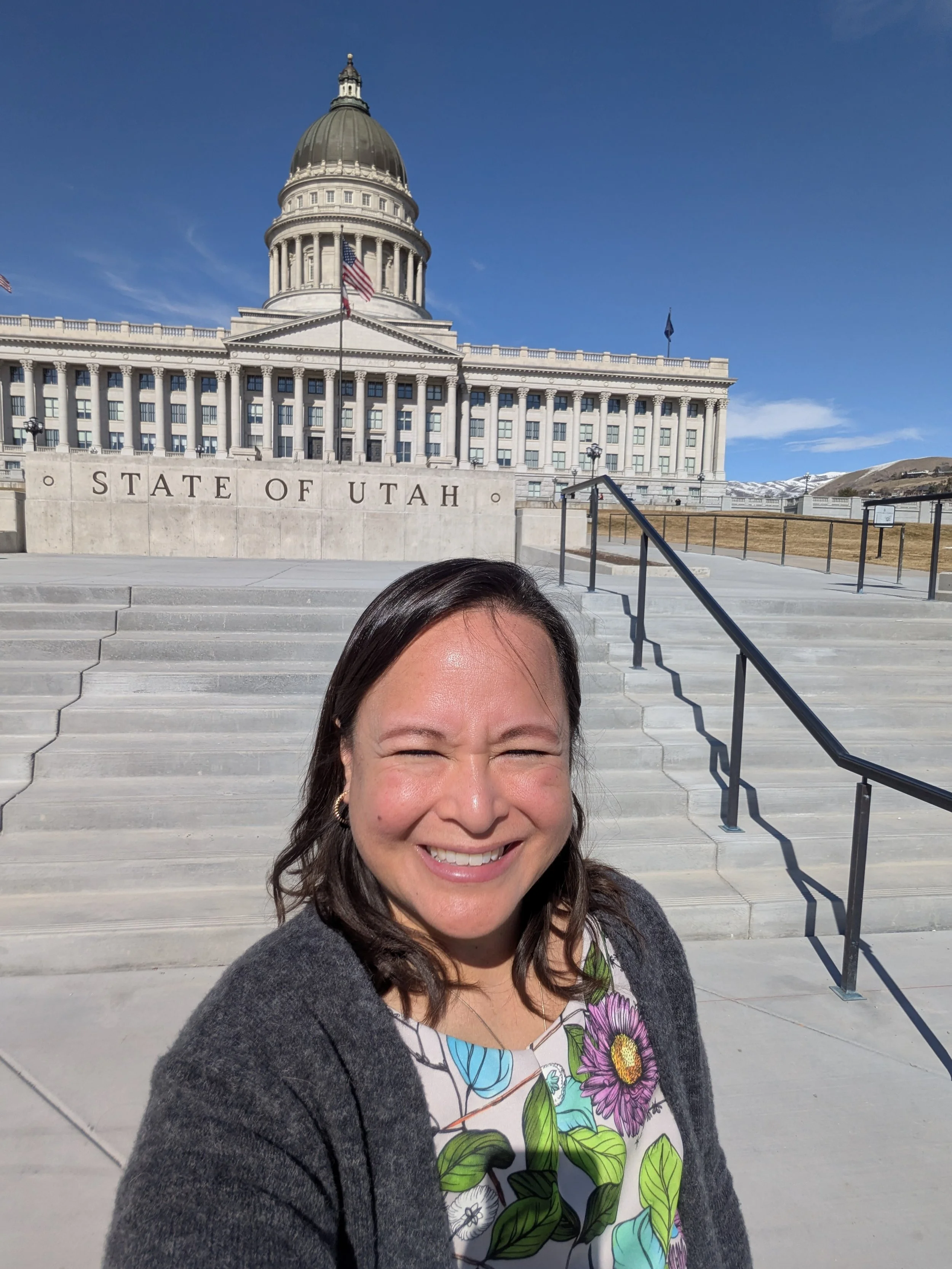 Smiling woman taking a selfie in front of the Utah State Capitol building with steps and a railing visible, clear blue sky in the background.