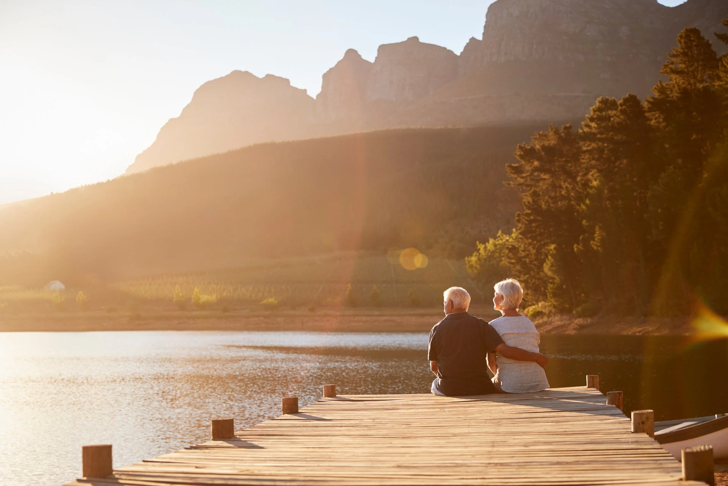 An elderly couple sitting together on a wooden dock by a lake during sunset, with mountains and trees in the background.