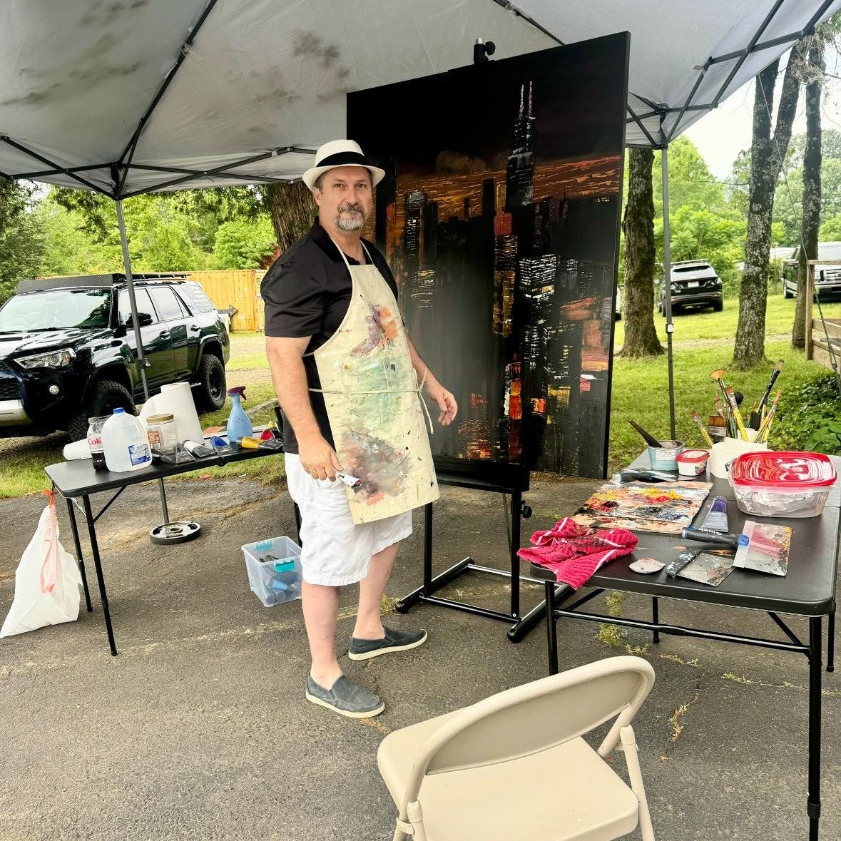An artist painting a city skyline on a large canvas outdoors under a canopy, with art supplies and tables around him.