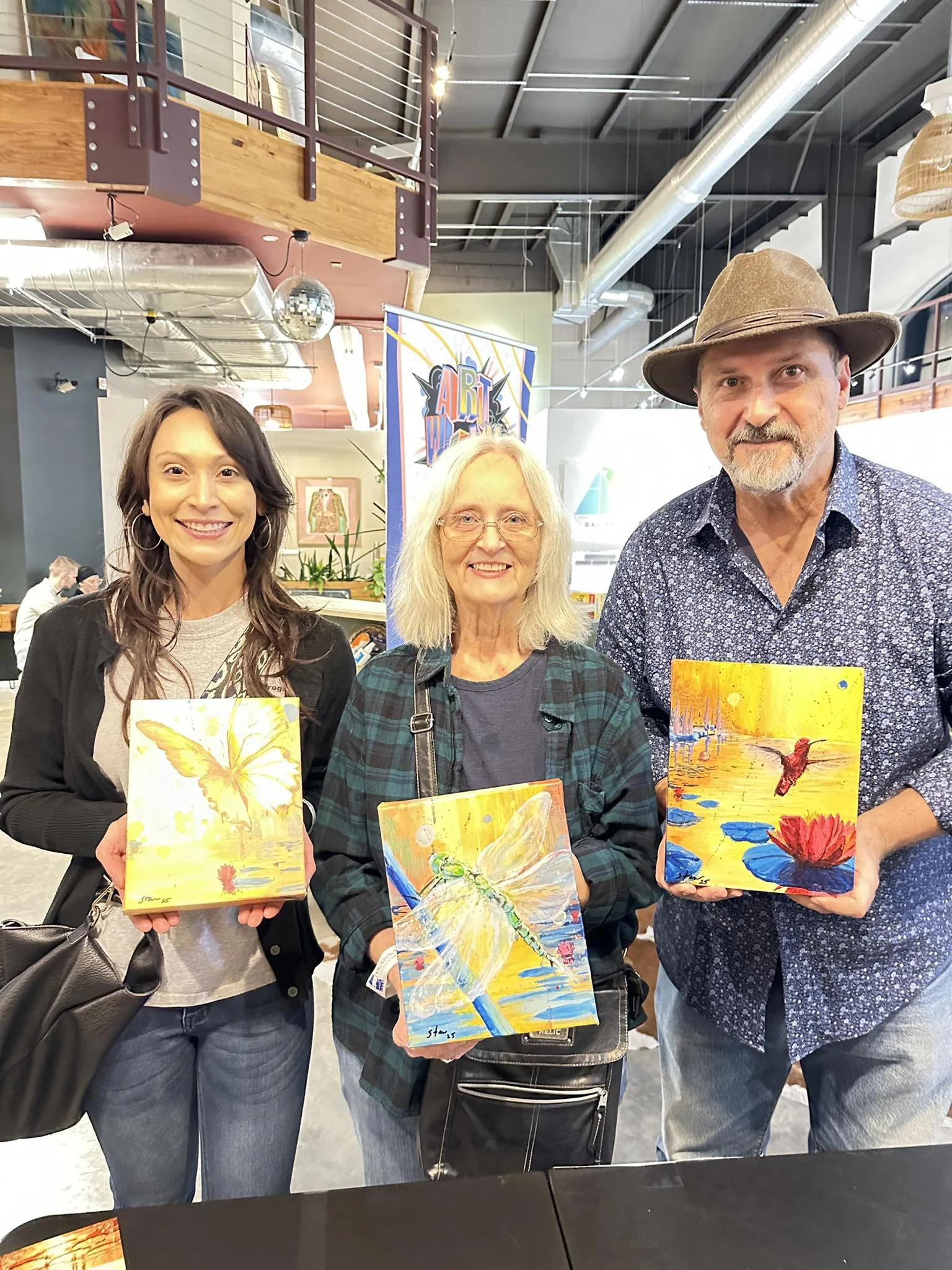Three people, two women and one man, smiling and holding small paintings of dragonflies by a body of water with a sunset background at an indoor art event.